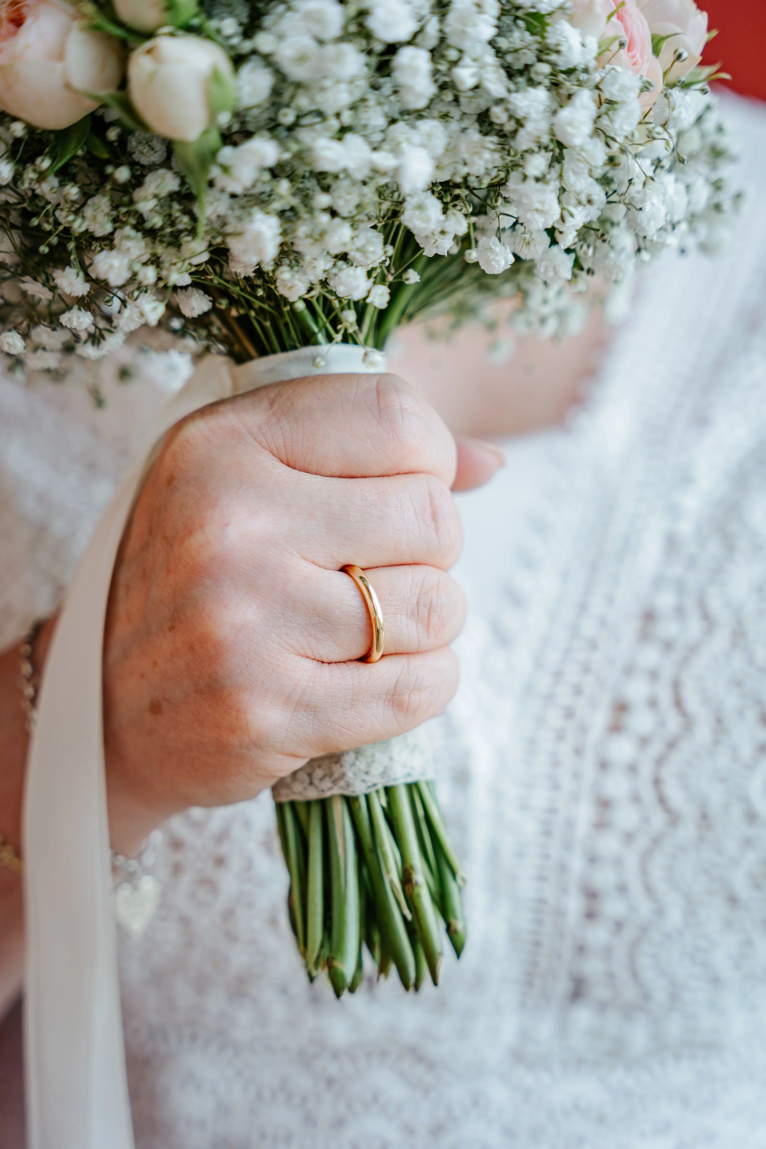 Nahaufnahme eines Brautpaares mit Blumenstrauß aus weißen Rosen und Schleierkraut; die Hände tragen goldene Eheringe.