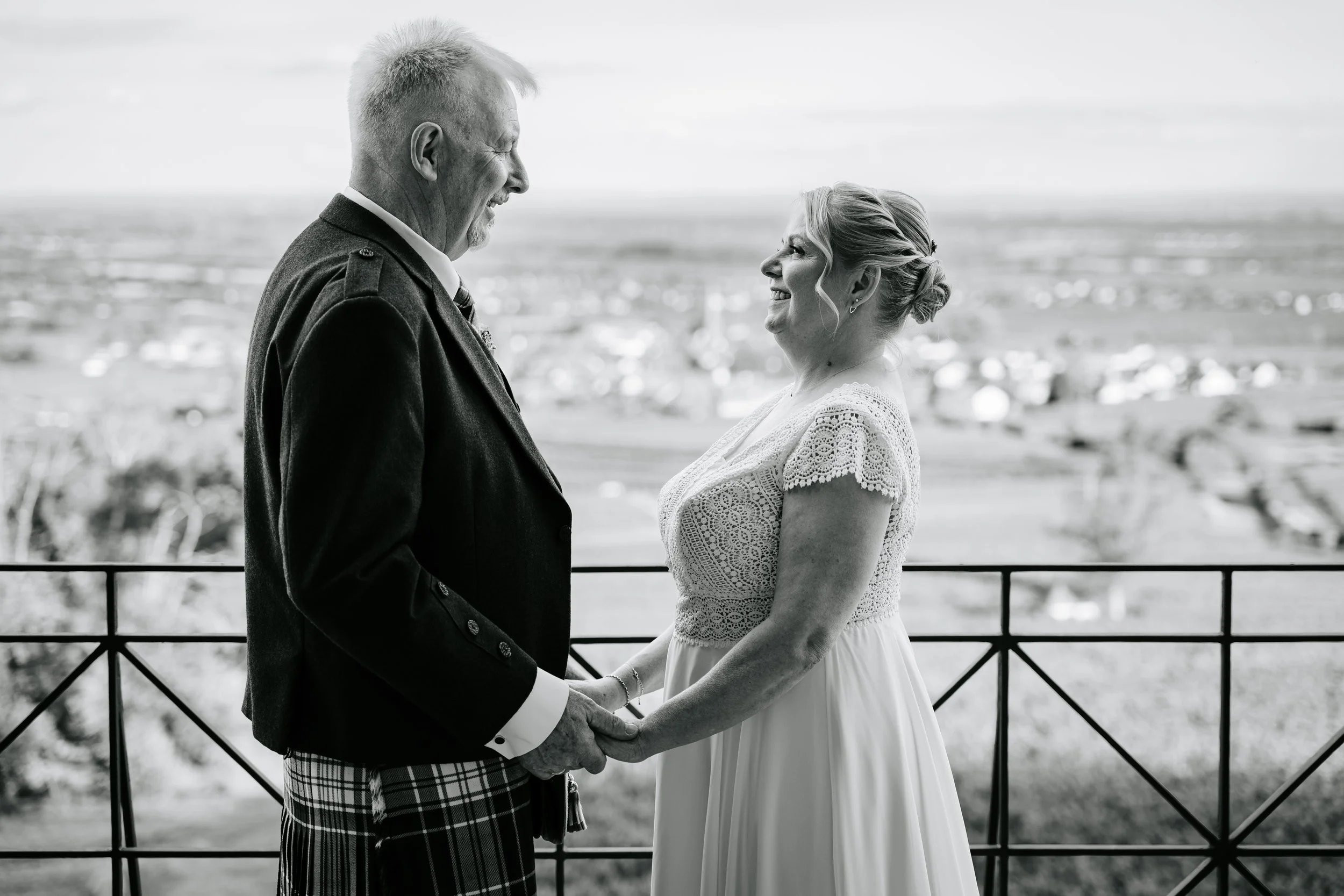 Schwarz-Weiß-Foto eines lächelnden Hochzeitspaars, das sich vor einer Küstenlandschaft an den Händen hält