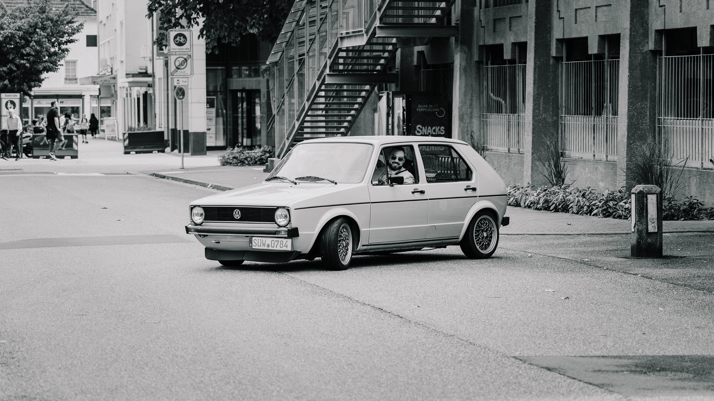 Schwarzweißfoto eines weißen Volkswagen Golf I mit Fahrer, geparkt in einer urbanen Straße vor einem modernen Gebäude.