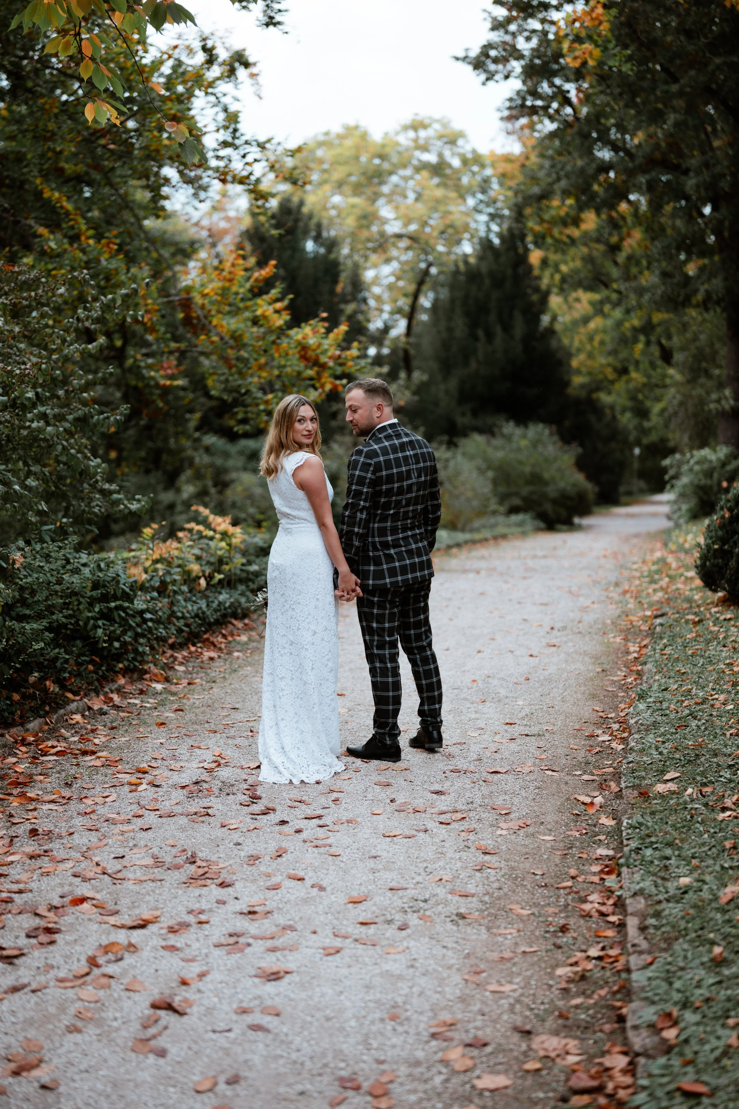 Ein Paar in formeller Kleidung hält sich an einem Spazierweg im Park während des Sonnenuntergangs, umgeben von herbstlichen Bäumen und gefallenen Blättern.
