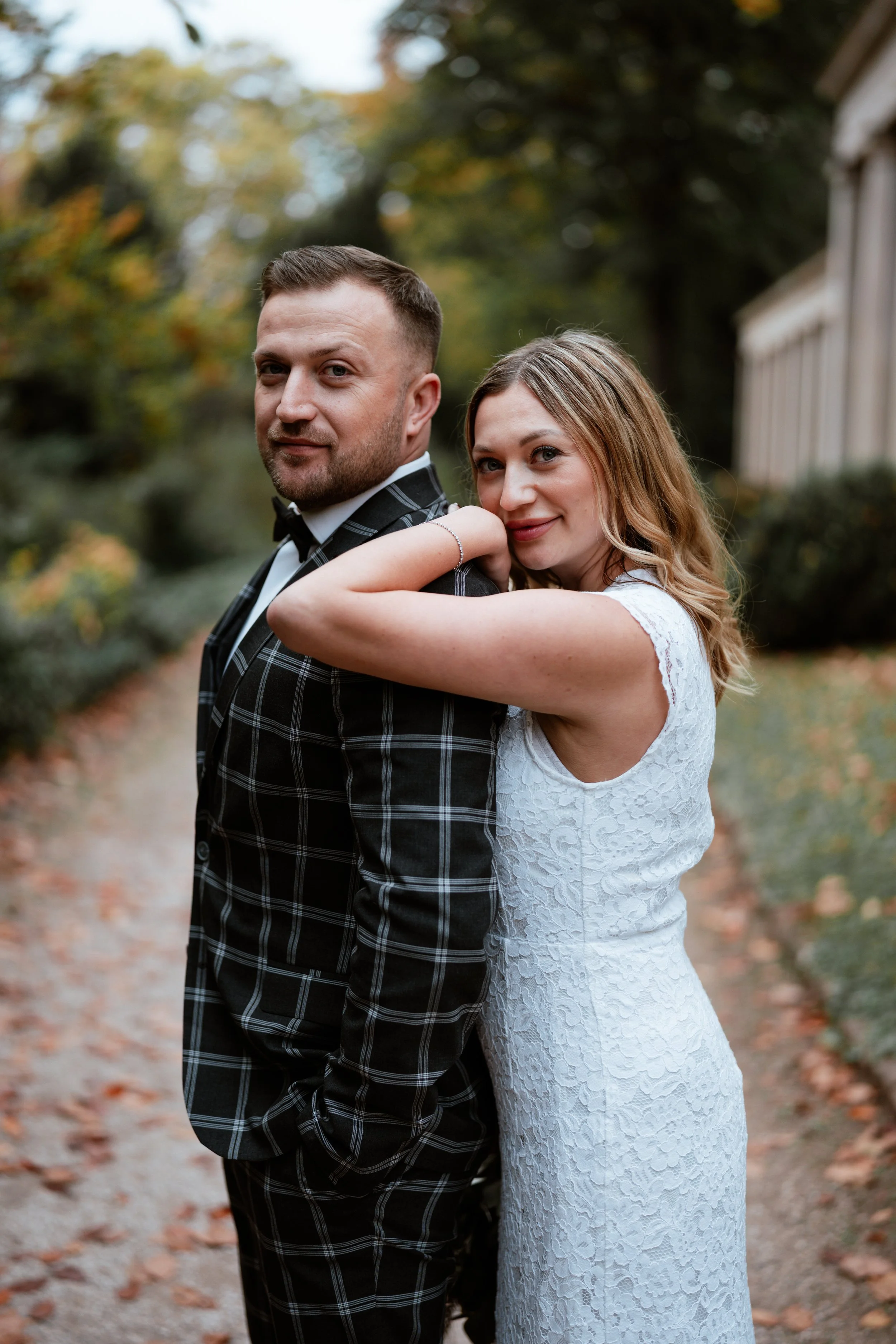 Ein Paar steht in einem gepflegten Gartenweg, umgeben von Bäumen mit herbstlichen Blättern, das Brautpaar vor einer Hochzeit trage ein elegantes Kleid und einen Anzug.