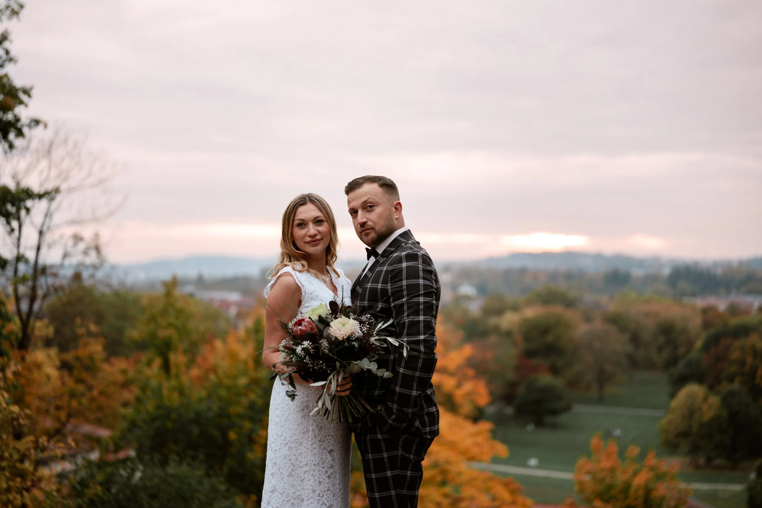 Ein Paar in Hochzeitskleidung steht zusammen auf einer Wiese mit buntem Herbstlaub, im Hintergrund ein bewölkter Himmel und eine Landschaft mit Bäumen.