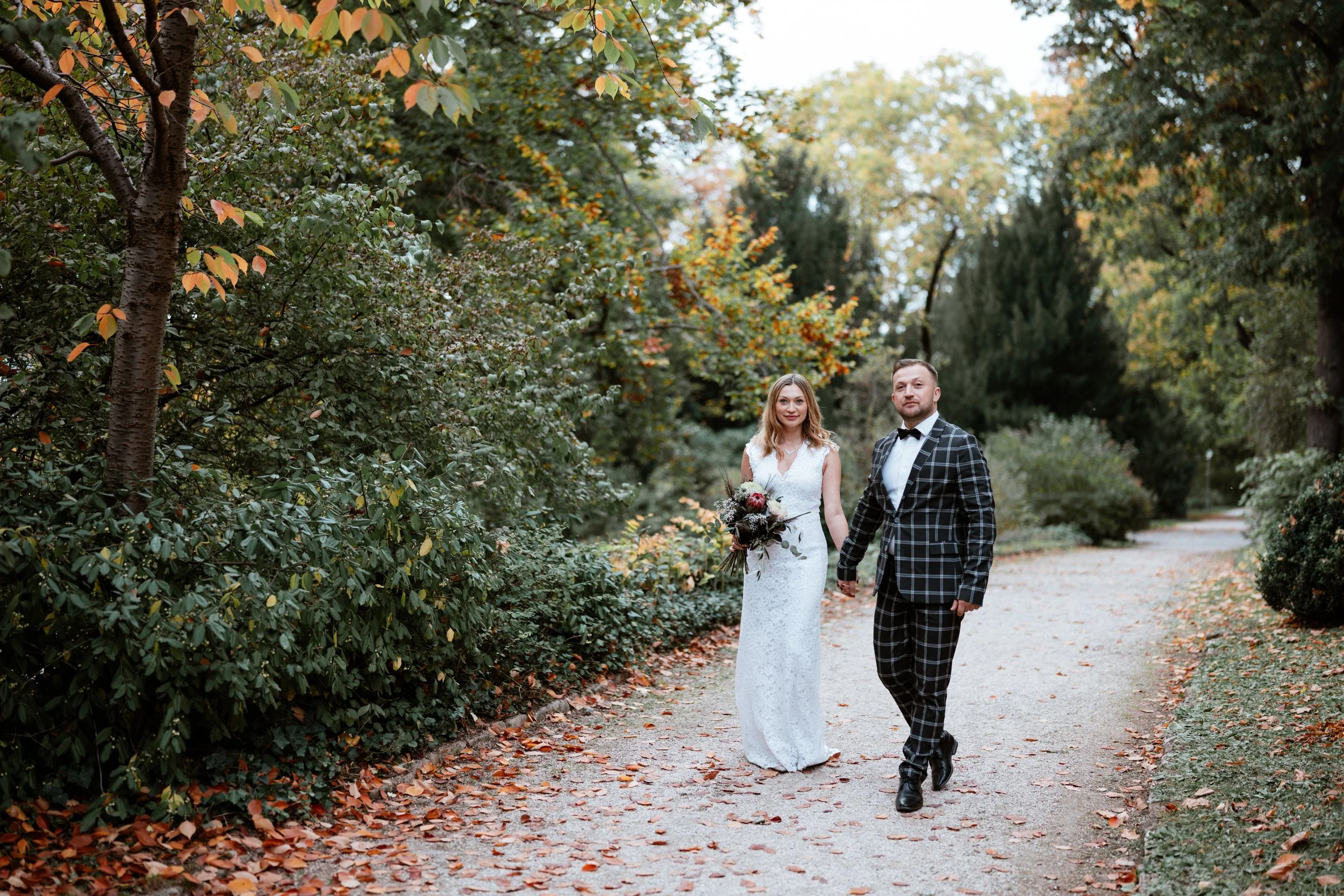 Ein Brautpaar in formeller Kleidung läuft in einem Park mit herbstlichen Bäumen und Laub