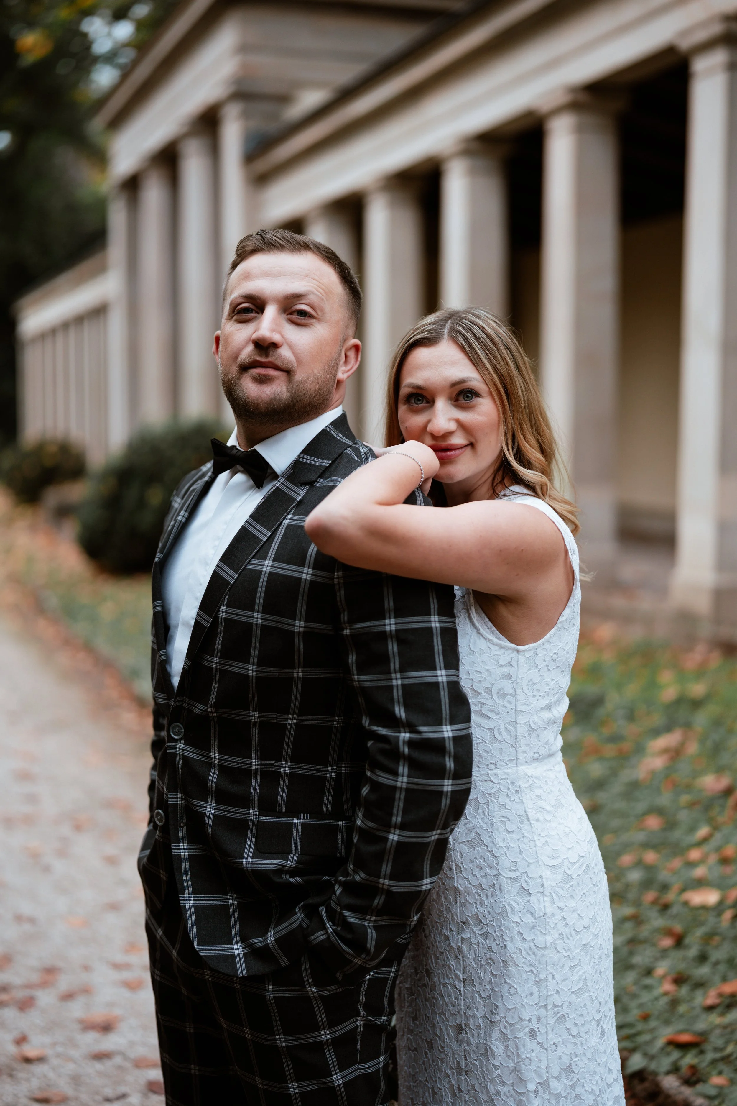 Ein Paar steht eng zusammen vor einer historischen Säulenstruktur im Freien, die Frau trägt ein weißes Hochzeitskleid und die Männer einen schwarzen Anzug mit Karo-Muster.