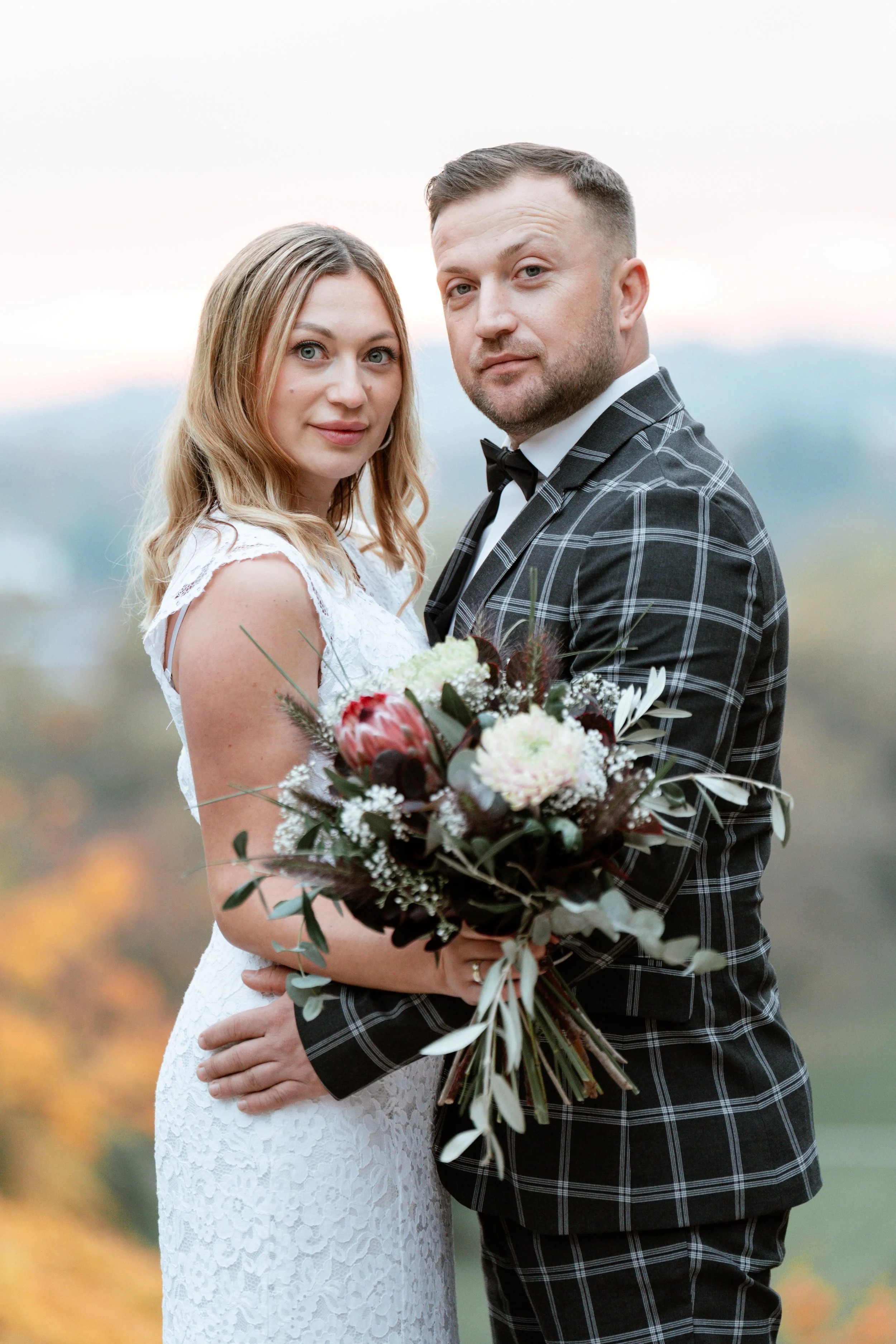 Ein Brautpaar steht im Freien, die Frau in einem weißen Hochzeitskleid und der Mann in einem schwarzen Kilt mit karierter Jacke, sie hält einen Brautstrauß.