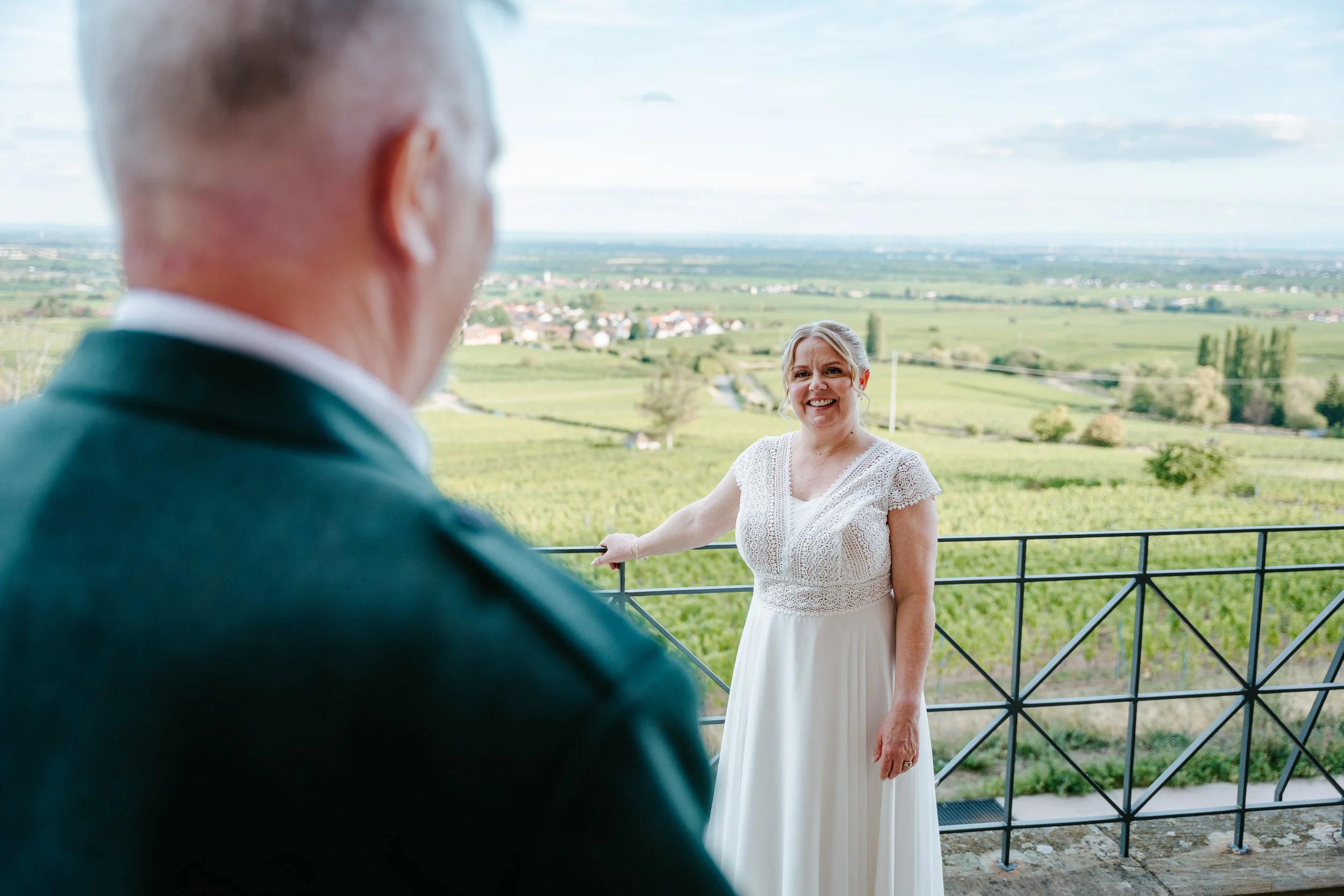 Lächelnde Frau im weißen Kleid auf einer Terrasse mit Weinberg- und Landschaftsblick, im Vordergrund ein Mann im Anzug.