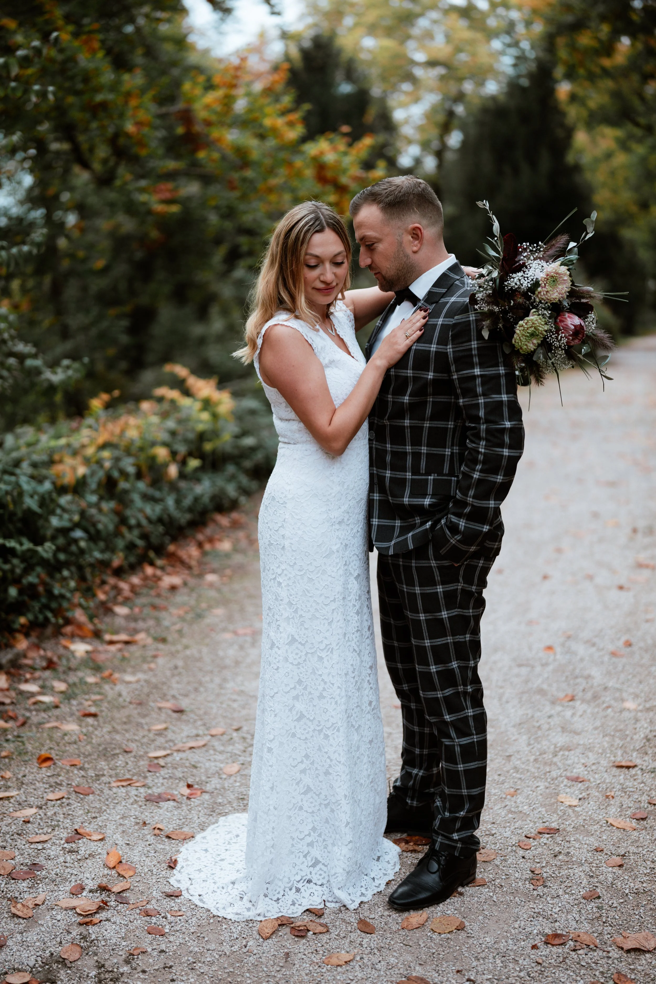 Ein Paar steht sich an einem Herbsttag in einem Park gegenüber, die Frau trägt ein weißes Hochzeitskleid und der Mann einen schwarzen Anzug mit Karo-Muster. Sie umarmen sich liebevoll, die Frau hält eine große Hochzeitsblumenstrauß auf den Rücken. Bl