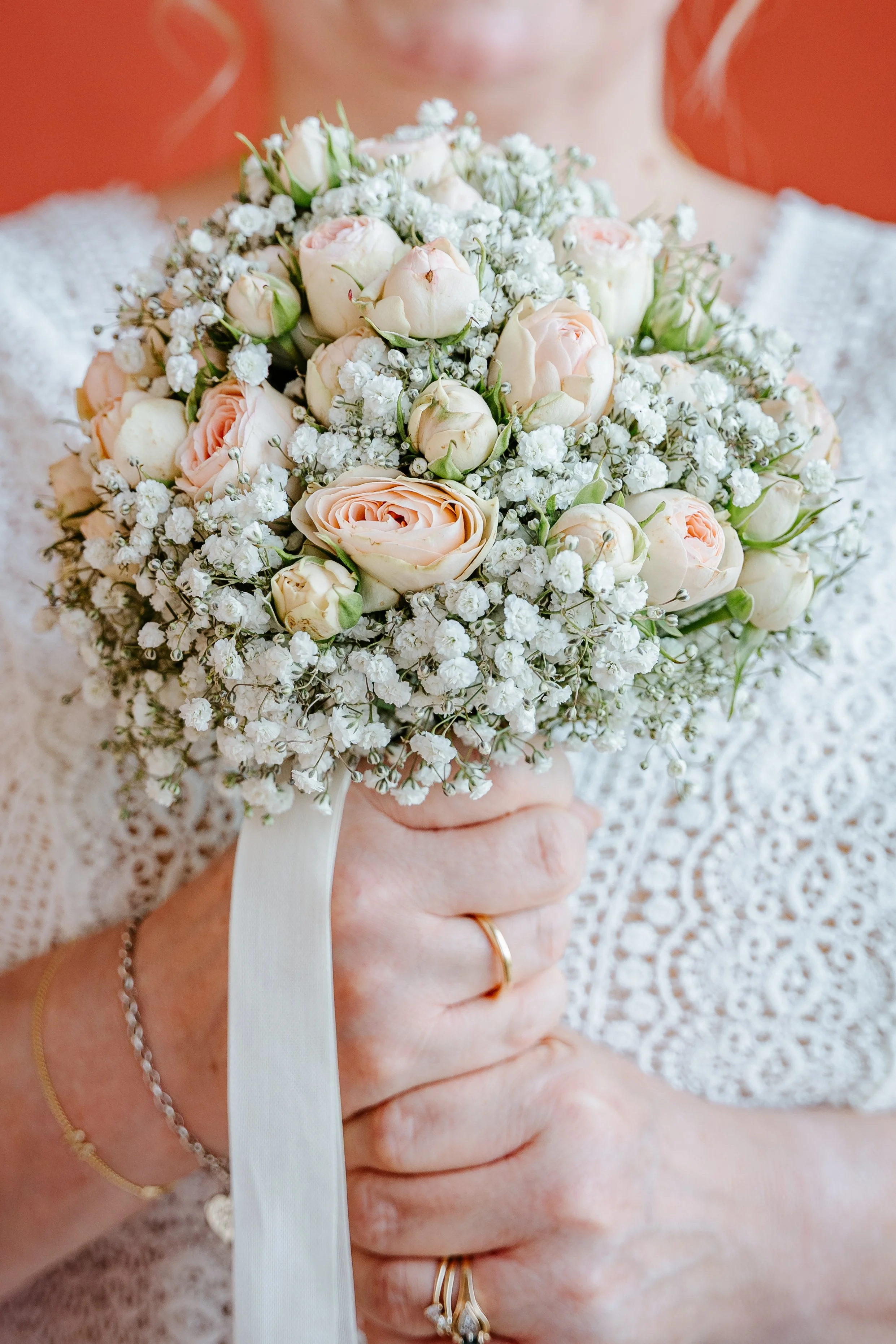 Nahaufnahme eines zarten Brautstraußes aus cremefarbenen Rosen, Ranunkeln und weißem Schleierkraut in den Händen einer Braut.
