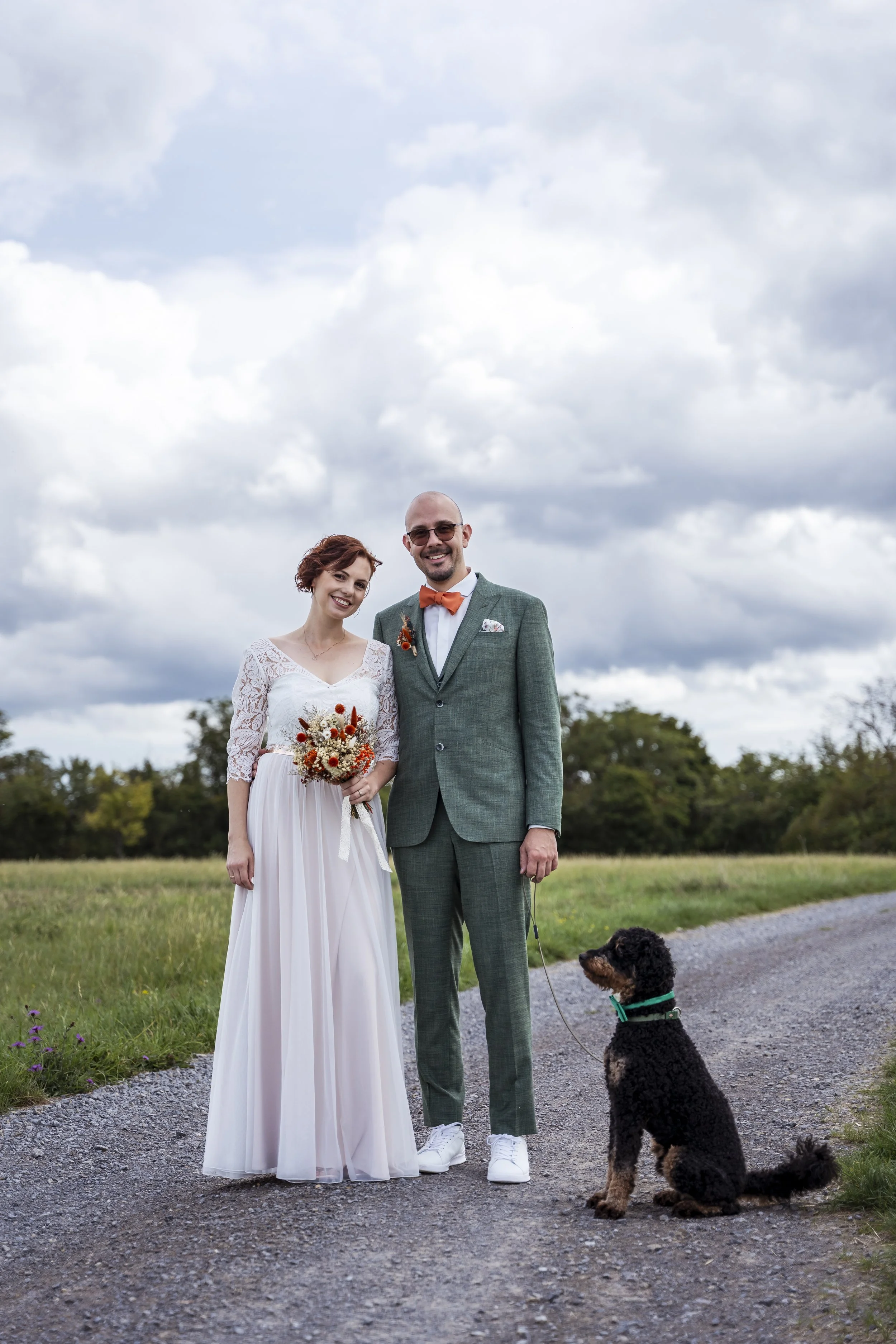 Ein Brautpaar steht im Freien auf einer landwirtschaftlichen Straße, die Frau trägt ein weißes Hochzeitskleid, der Mann einen grünen Anzug mit Orangenen Akzenten, und ein Hund sitzt neben ihnen, während der Himmel bewölkt ist.