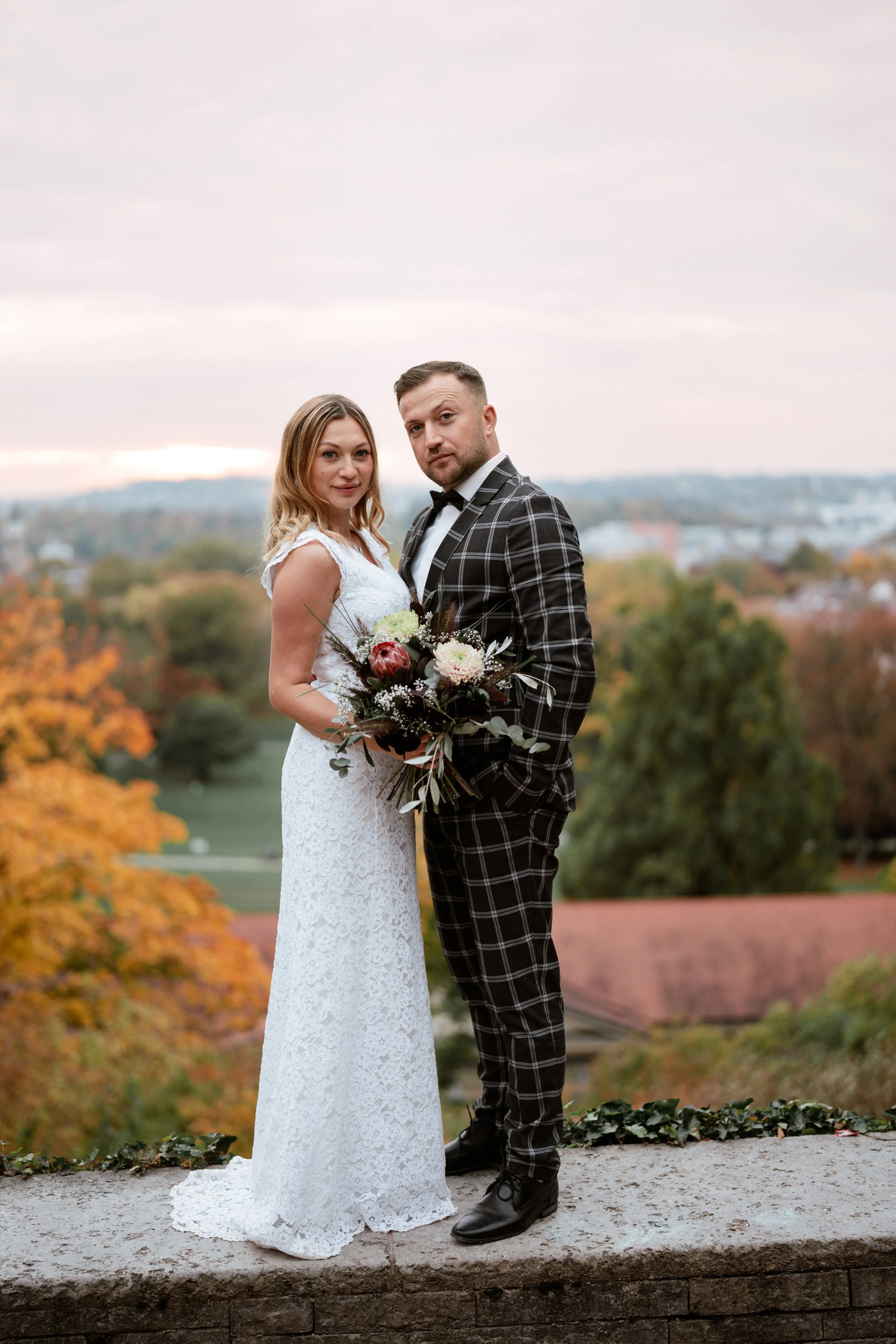 Ein Hochzeitsfoto eines Paares, die Frau trägt ein weißes Hochzeitskleid und hält einen Blumenstrauß, der Mann trägt einen schwarzen Anzug mit weißem Hemd und Fliege, im Hintergrund sind Herbstbäume und eine Landschaft sichtbar.