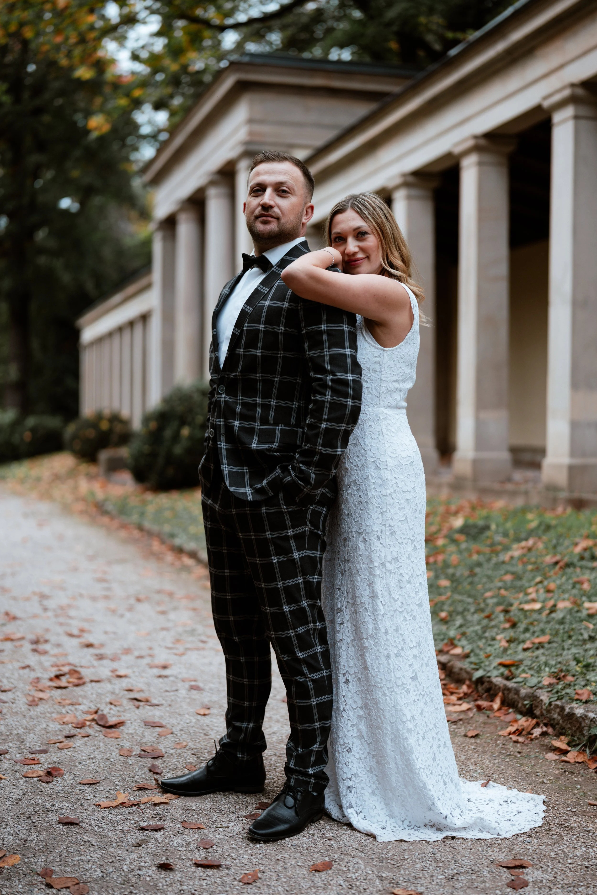 Ein Brautpaar in formeller Kleidung, draußen vor einem Gebäude mit Säulen, umgeben von Herbstlaub.
