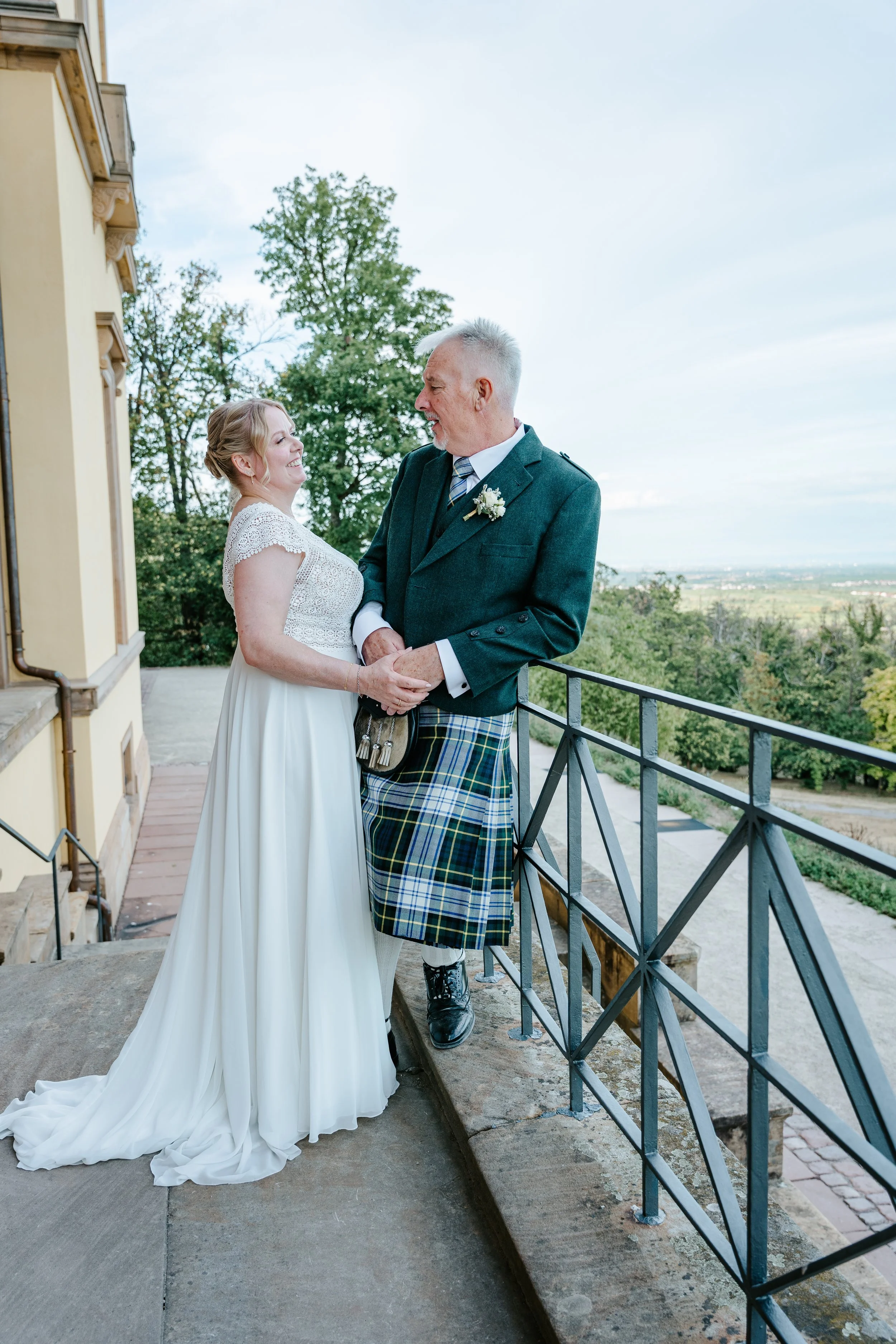 Brautpaar auf einer Terrasse: Die Braut im weißen Spitzenkleid lächelt den Bräutigam im grünen Anzug und Schottenrock an.