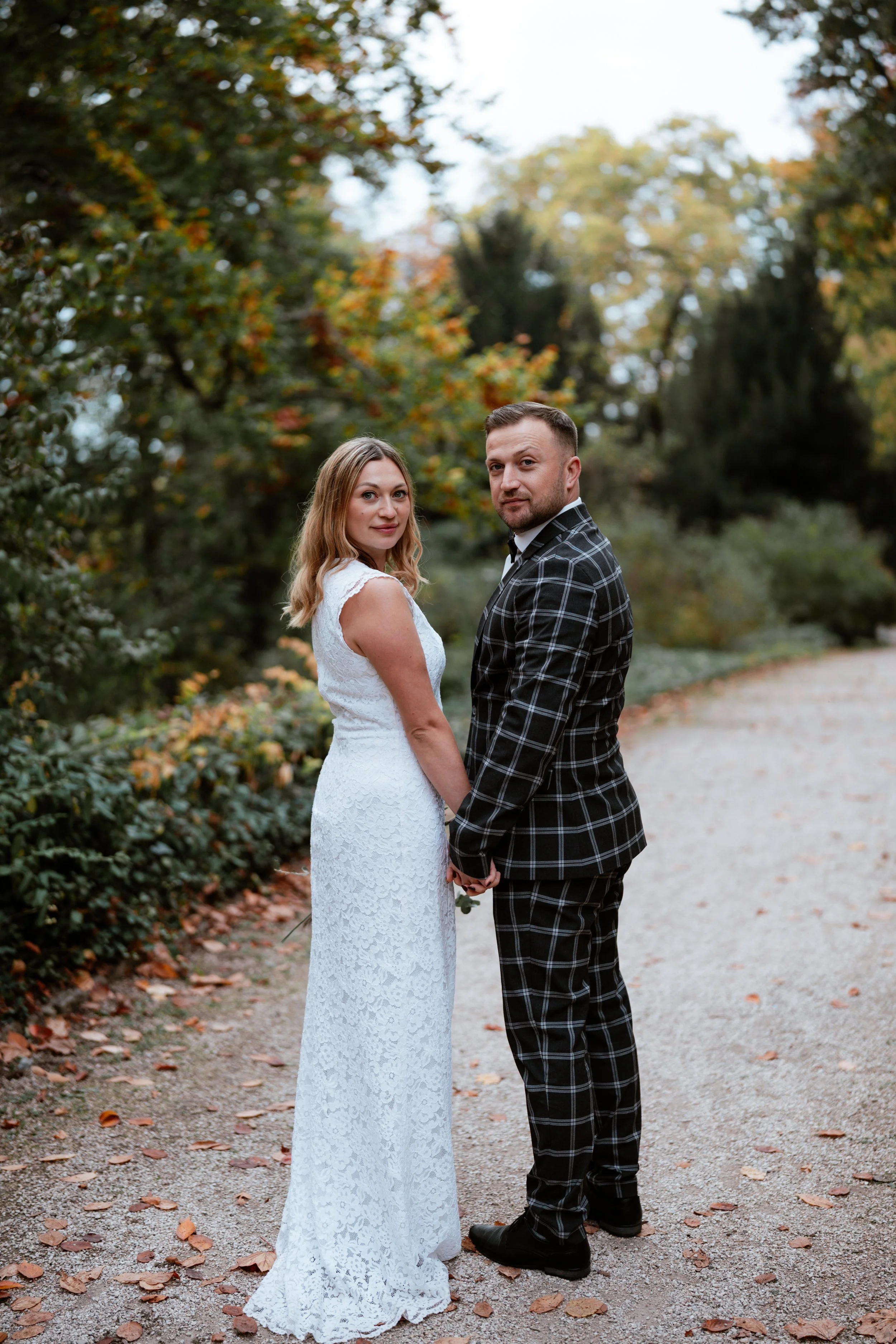 Ein Paar in Hochzeitskleidung hält sich an einem Weg in der Natur, umgeben von Bäumen mit herbstlichen Farben, bei Sonnenuntergang.