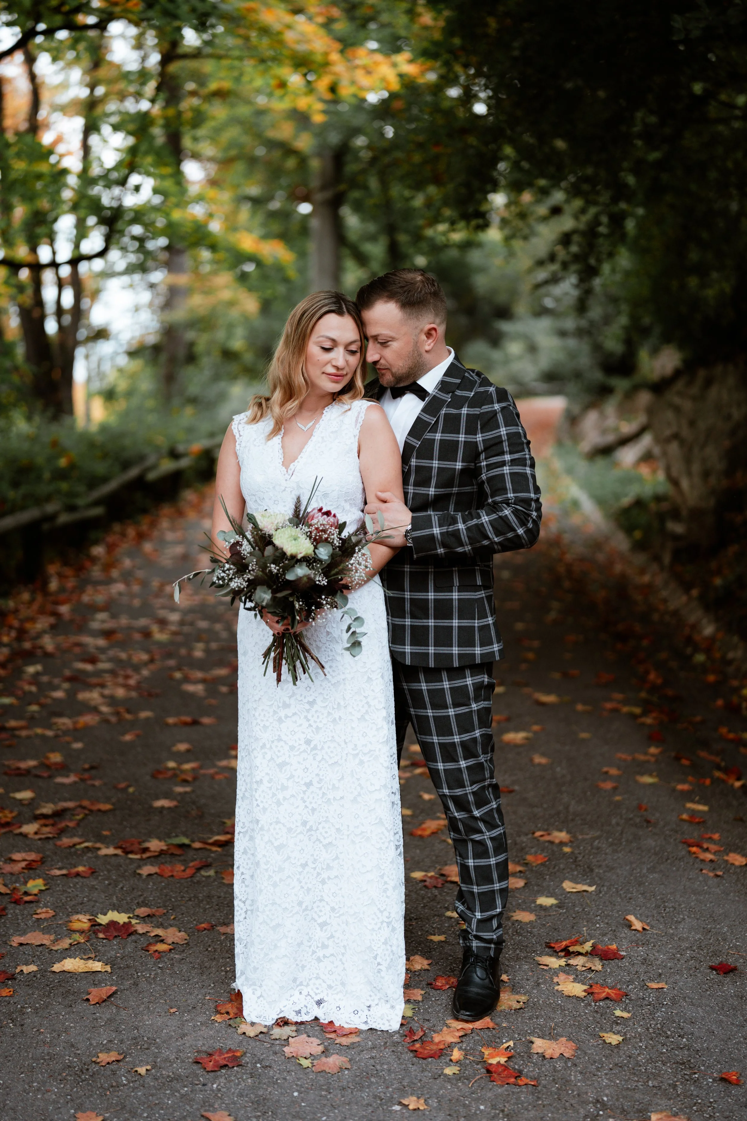 Ein Brautpaar im Wald an einem Herbsttag, die Braut trägt ein langes weißes Kleid und hält einen Blumenstrauß, der Bräutigam trägt ein schwarz-weiß kariertes Anzug mit Fliege.