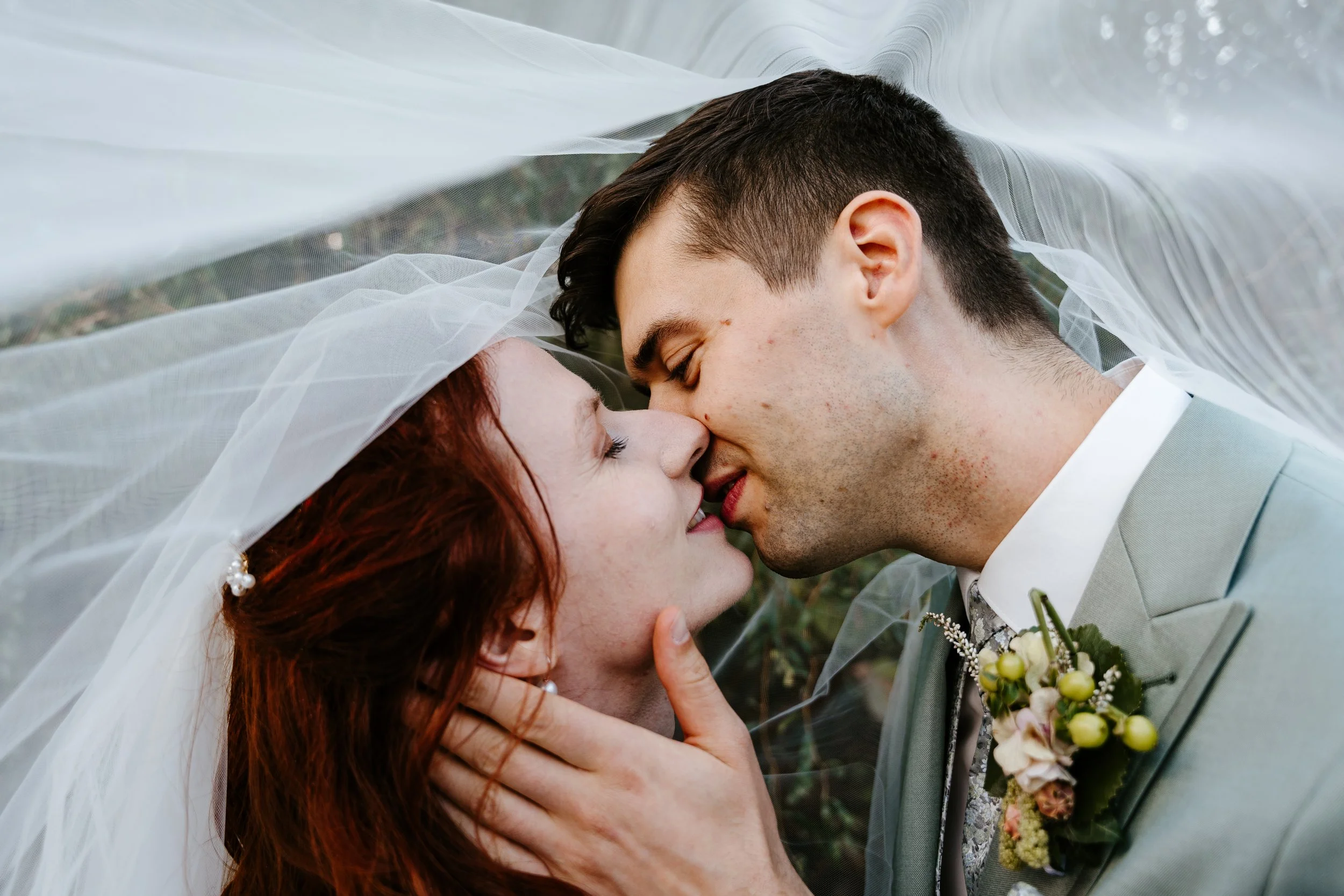 Brautpaar küsst sich unter einem weißen Hochzeitsschleier; der Bräutigam hält einen Blumenstrauß.