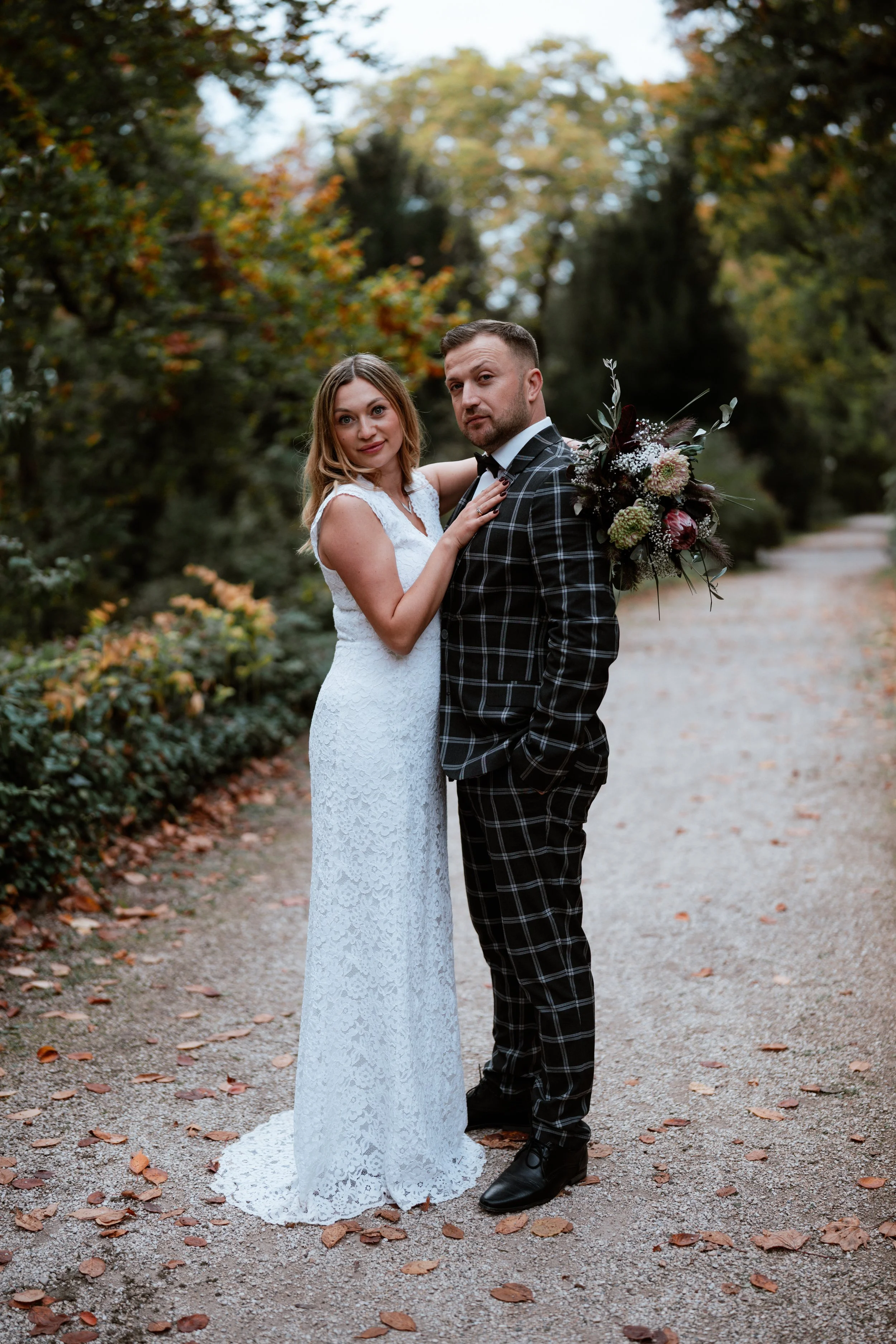 Ein Brautpaar steht auf einem abgelegenen Weg in einem bewachsenen Park mit herbstlichen Blättern auf dem Boden. Die Braut trägt ein weißes Spitzenkleid, der Bräutigam einen schwarzen Anzug mit Karo-Muster und hält einen großen Blumenstrauß. Im Hinte