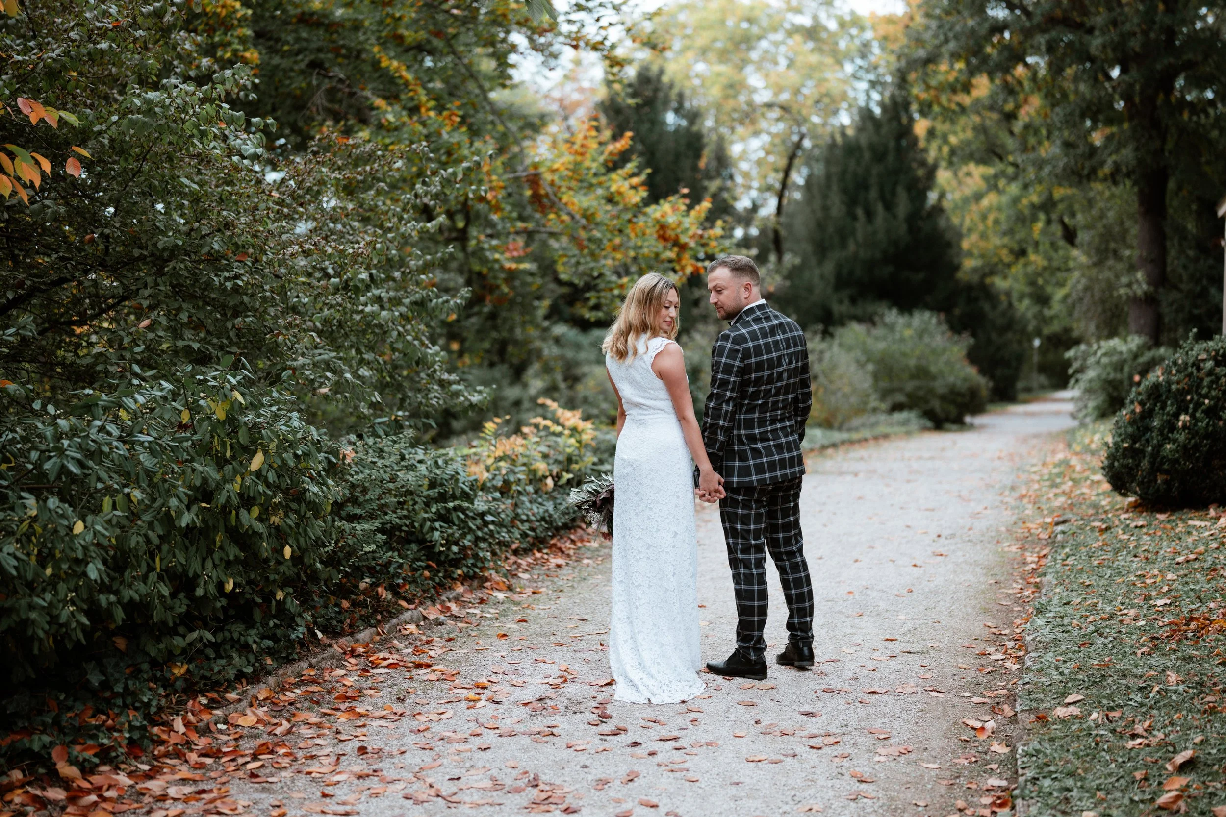 Ein Paar hält Händchen auf einem gepflasterten Weg, umgeben von herbstlicher Vegetation, vermutlich während eines Fotoshootings für eine Hochzeit.