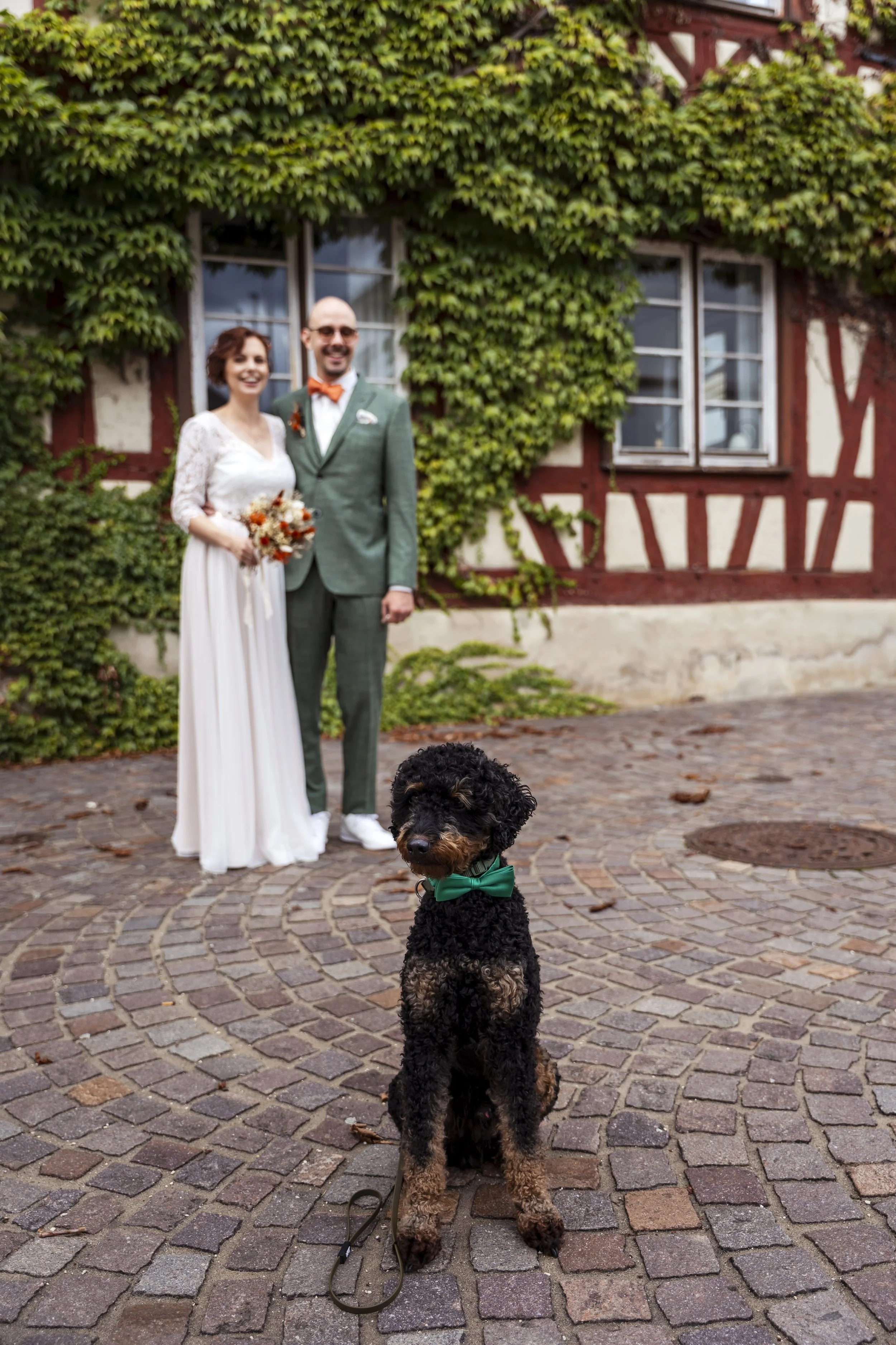 Ein Brautpaar steht auf einem gepflasterten Platz vor einem Haus mit Efeu. Im Vordergrund sitzt ein schwarzer Hund mit braunen Flecken, trägt eine grüne Fliege, und ist an einer Leine.