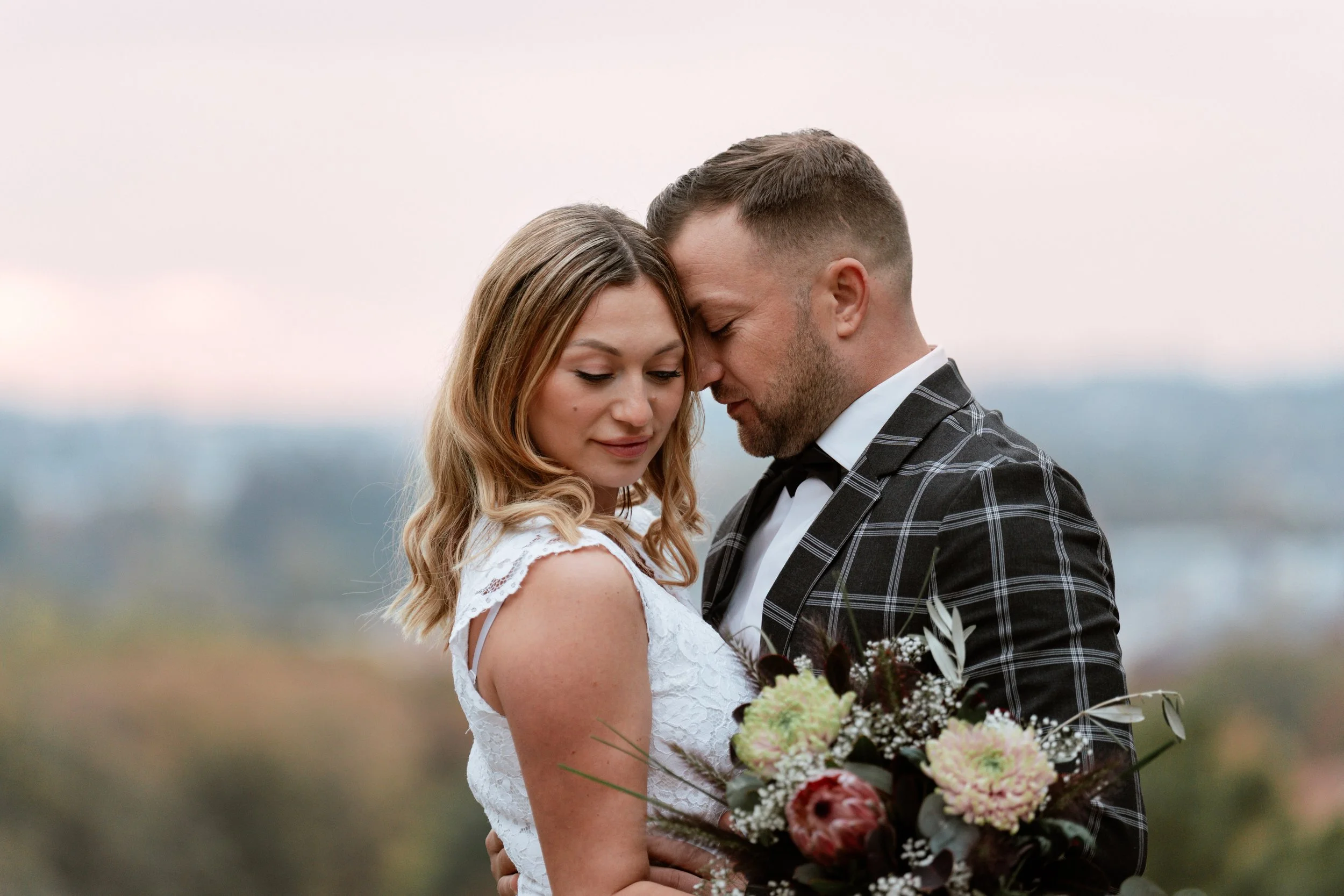 Ein verliebtes Paar bei einer Hochzeit, die sich liebevoll umarmen, mit einem Blumenstrauß in der Hand. Der Himmel ist bewölkt.