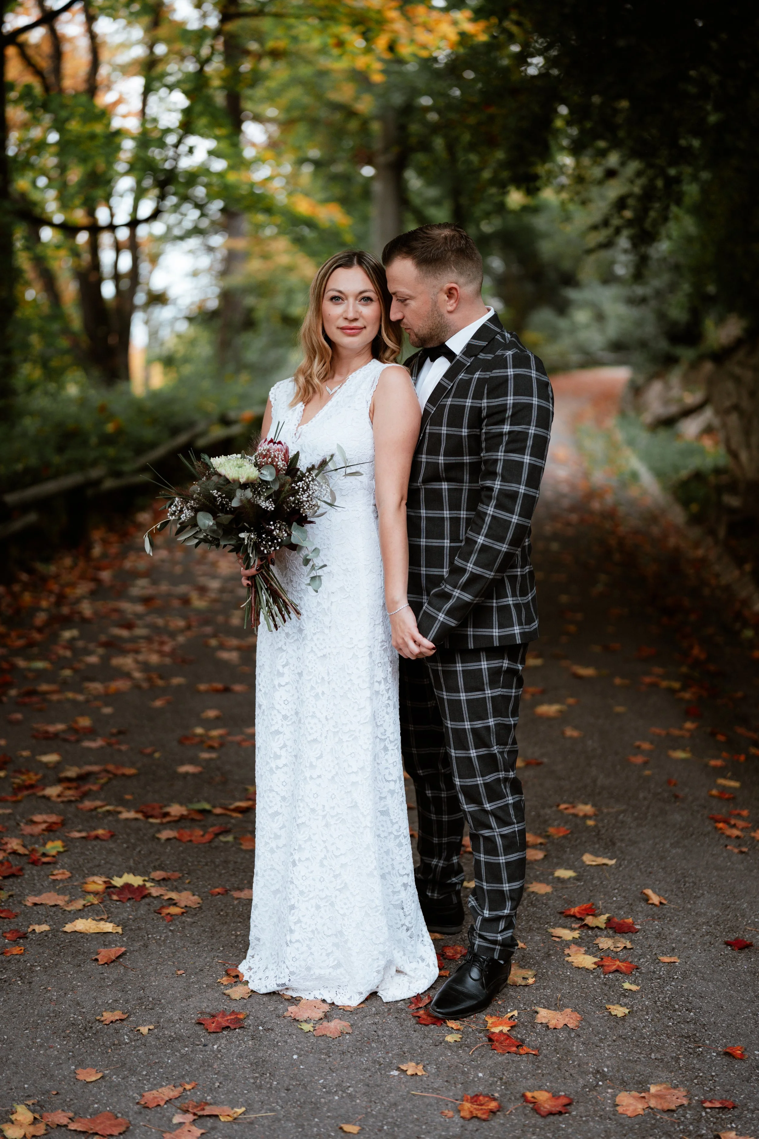 Ein Brautpaar steht im Herbst in einem Waldweg. Die Braut trägt ein weißes Hochzeitskleid und hält einen Blumenstrauß. Der Bräutigam trägt einen schwarz-weiß karierten Anzug. Die Blätter auf dem Boden sind rot, orange und gelb.