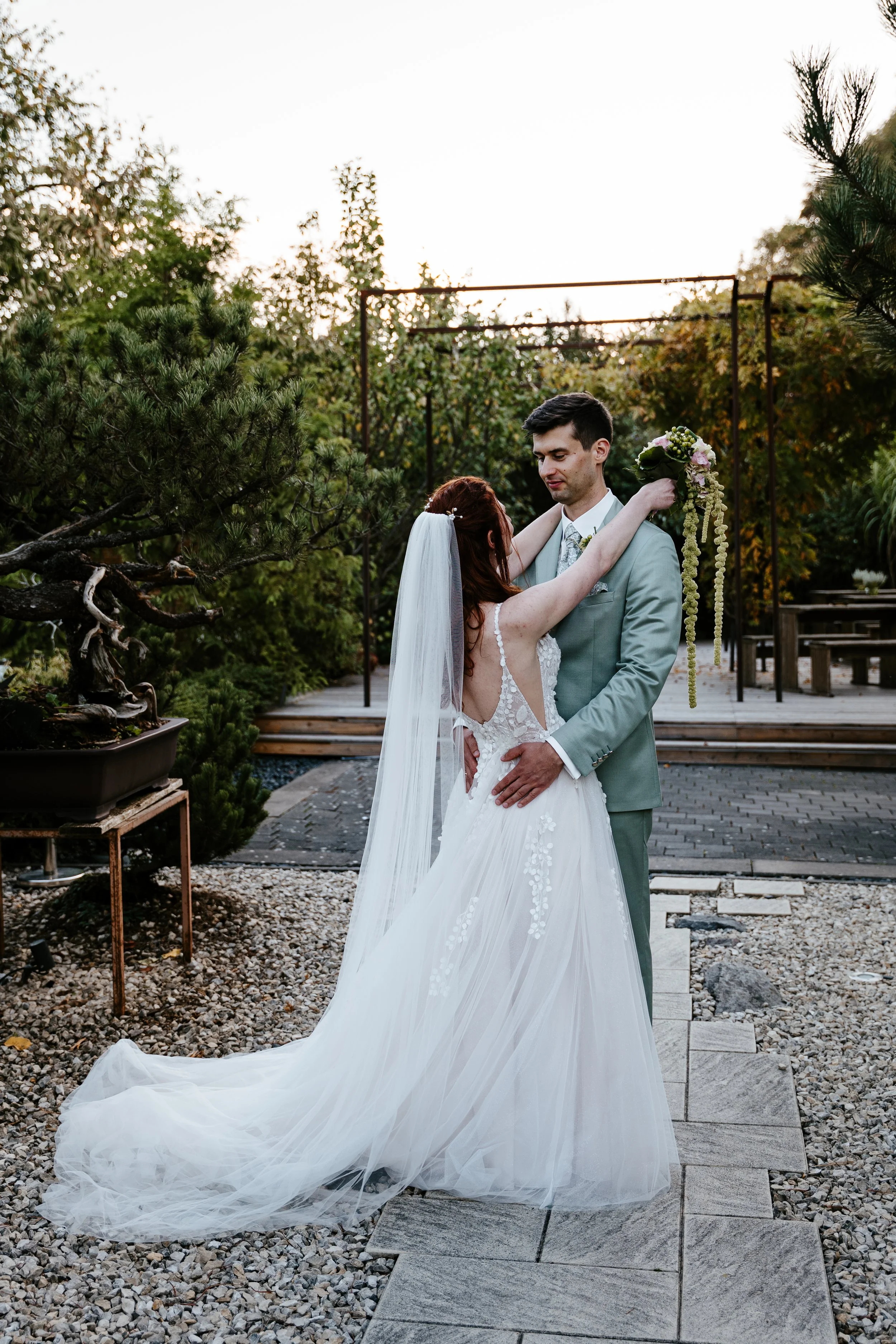 Brautpaar umarmt sich im botanischen Garten; die Braut trägt ein langes Spitzenkleid mit Schleier und hält einen Blumenstrauß.