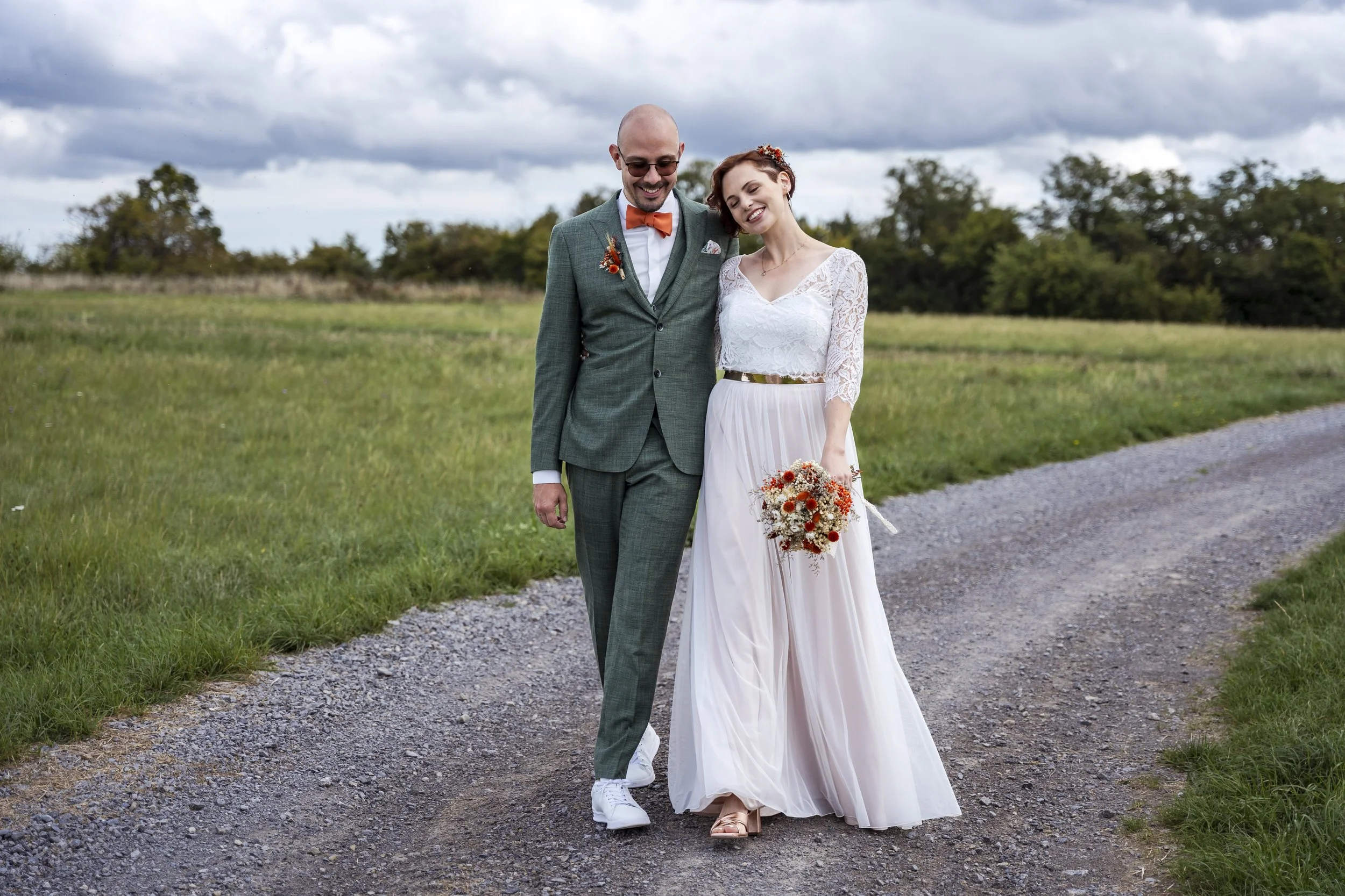 Ein Brautpaar spaziert lachend auf einem Weg in einer ländlichen Gegend, die Braut trägt ein weißes Hochzeitskleid mit einem Blumenbouquet, der Bräutigam trägt einen grünen Anzug mit orangefarbener Fliege.