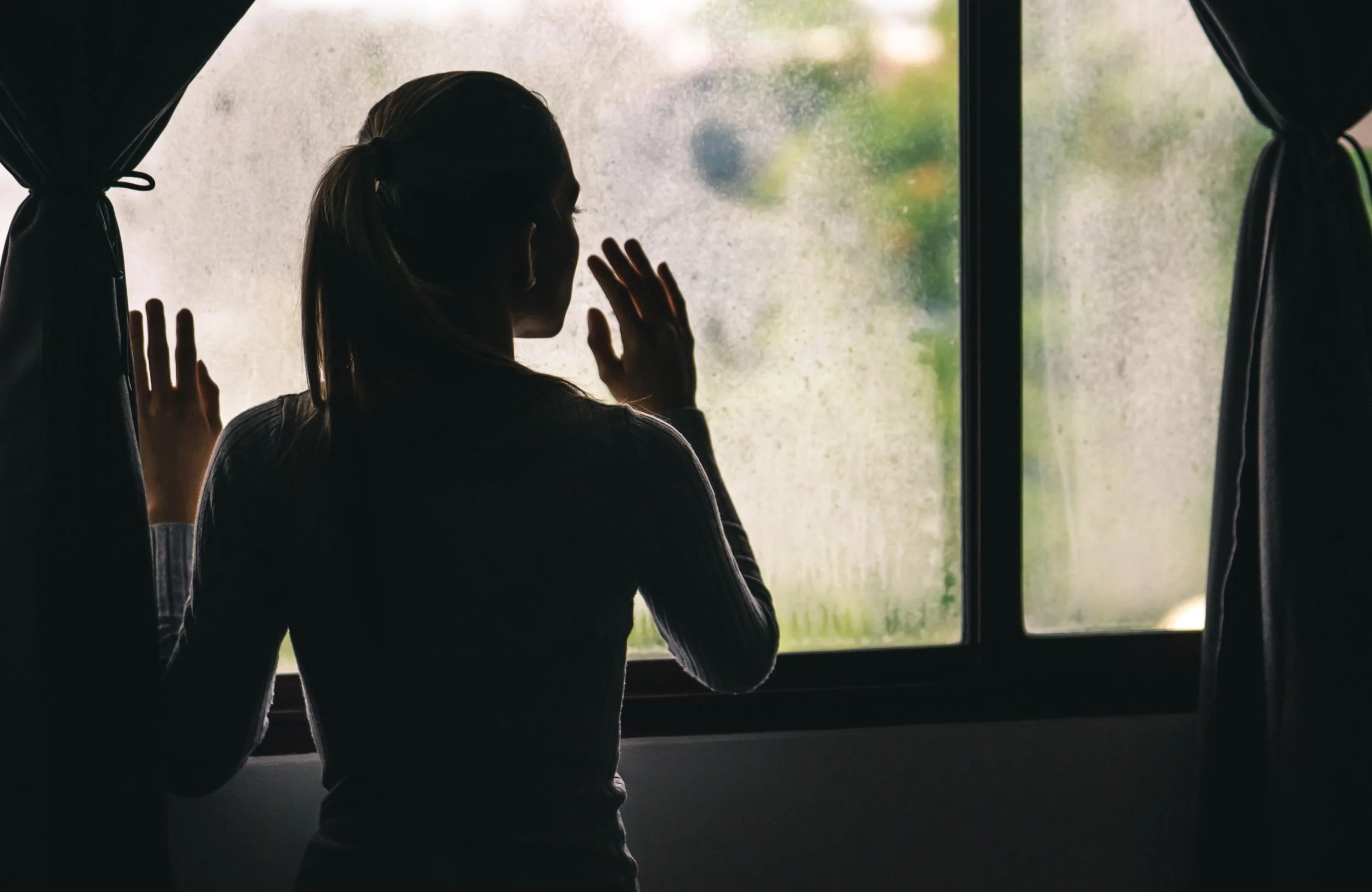 Eine Frau steht vor einem Fenster, berührt es leicht mit den Händen und schaut nach draußen. Die Scheibe ist verschwommen, der Hintergrund ist unscharf mit Grün und Himmel zu erkennen.