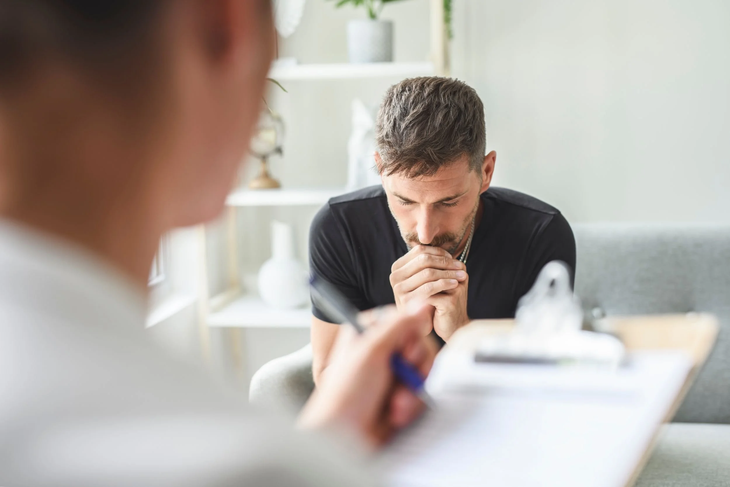 Ein Mann sitzt nachdenklich an einem Tisch in einem hellen Raum, während eine andere Person im Vordergrund dessen Gesicht und Aktivitäten unscharf sind.
