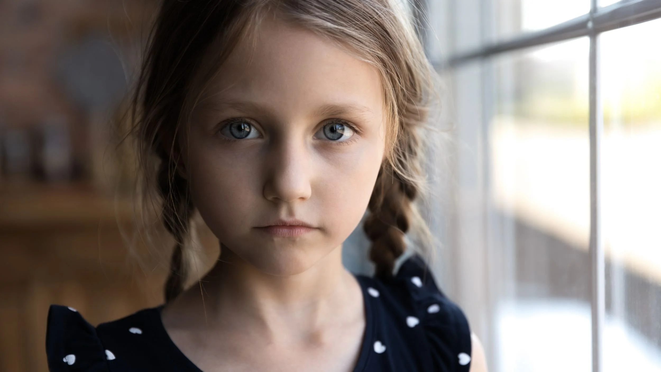 Nahaufnahme eines Mädchens mit blauen Augen und braunen, lockigen Haaren, die an einer Fensterseite steht.