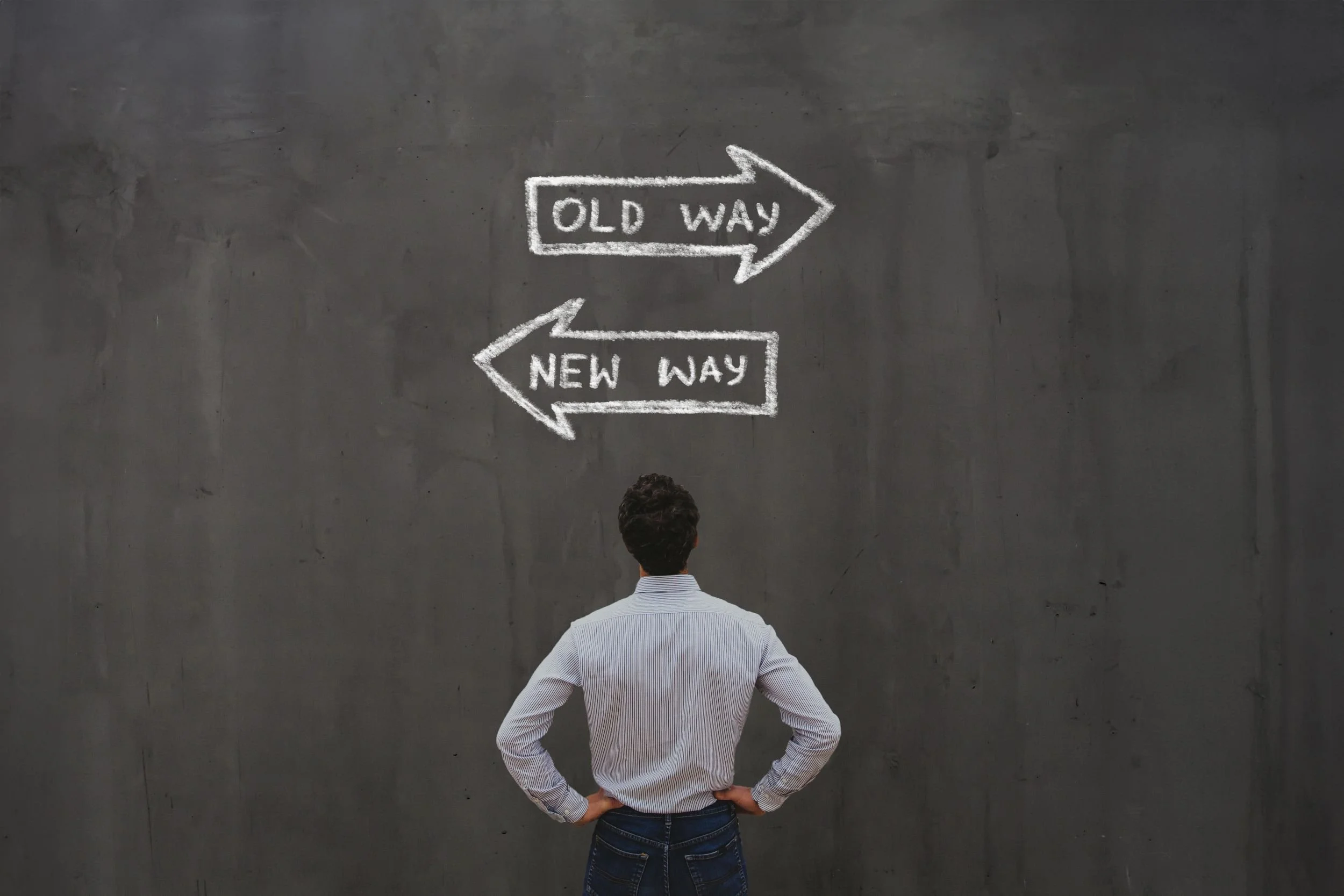 Ein Mann steht vor einer schwarzen Wand mit zwei Handgeschriebenen Pfeilen in Schulkreide, die 'OLD WAY' nach rechts und 'NEW WAY' nach links zeigen.