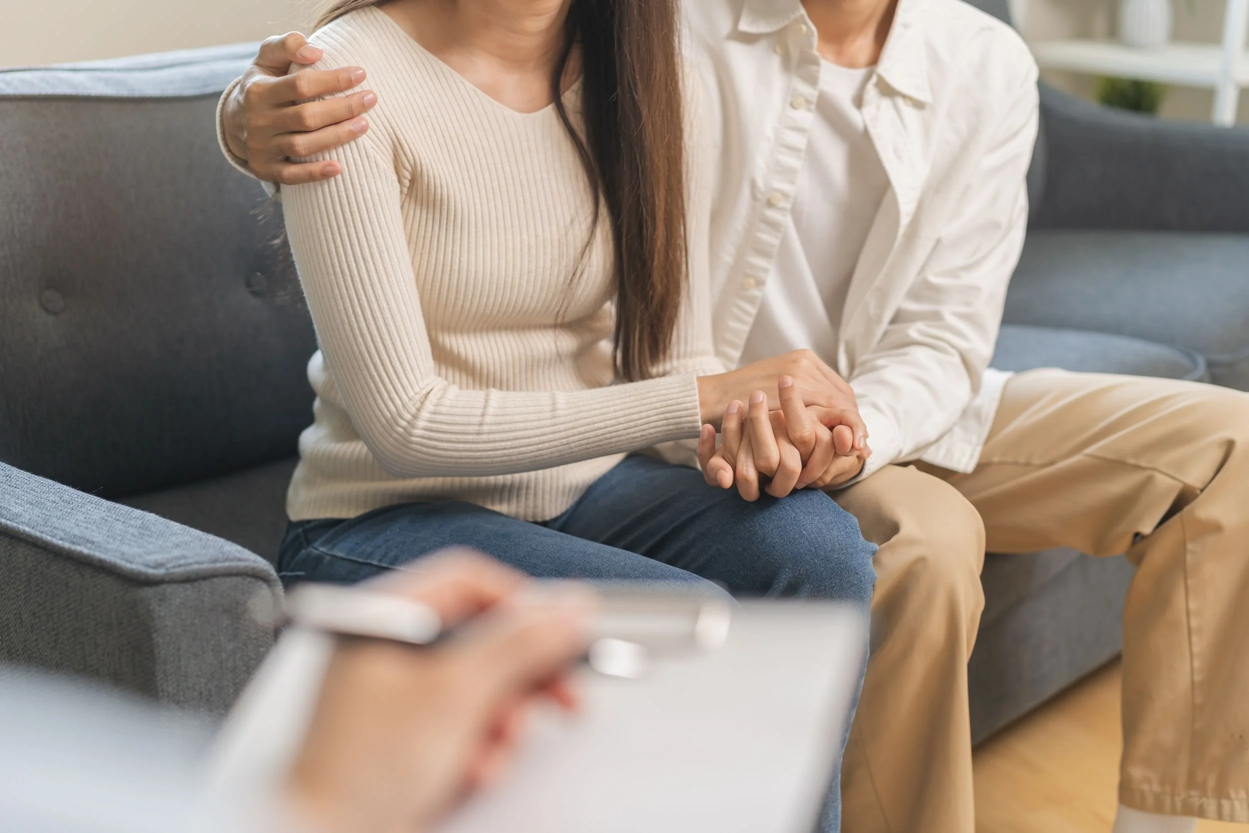 Zwei Menschen sitzen in einem Wohnzimmer, eine Person berührt die Schulter der anderen Person.