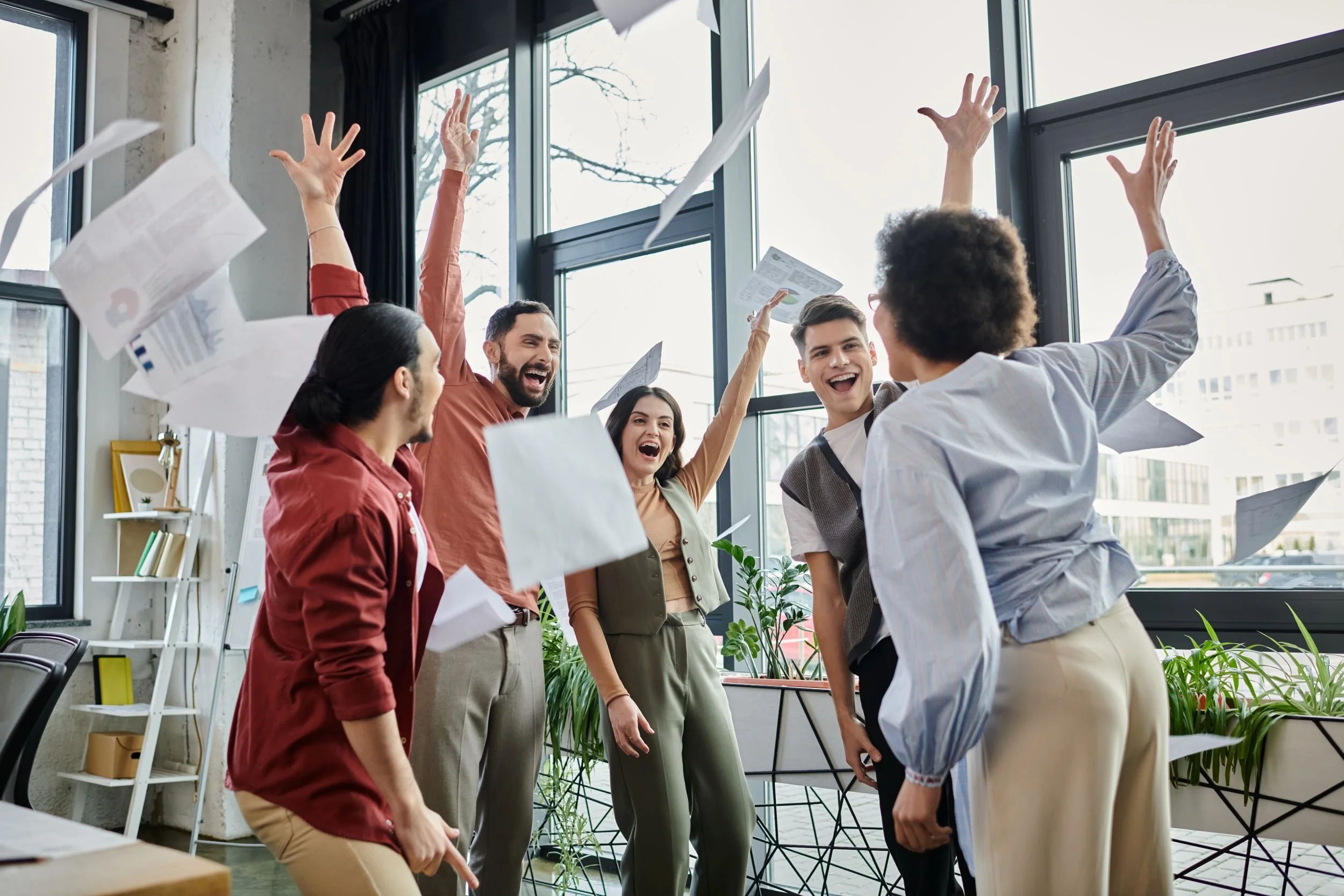 Grupp von Menschen in einem Büro, die jubeln und applaudieren, während Papierflugzeuge fliegen.