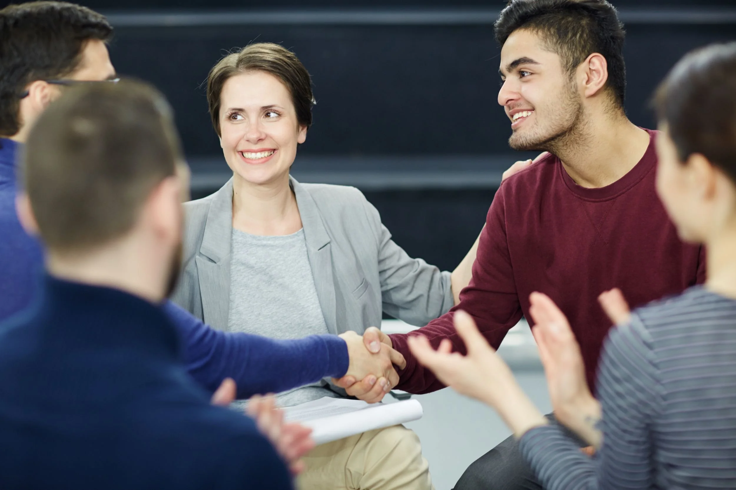 Menschen, die in einem Kreis sitzen, begrüßen sich mit Handschlägen und unterhalten sich in einem Raum.