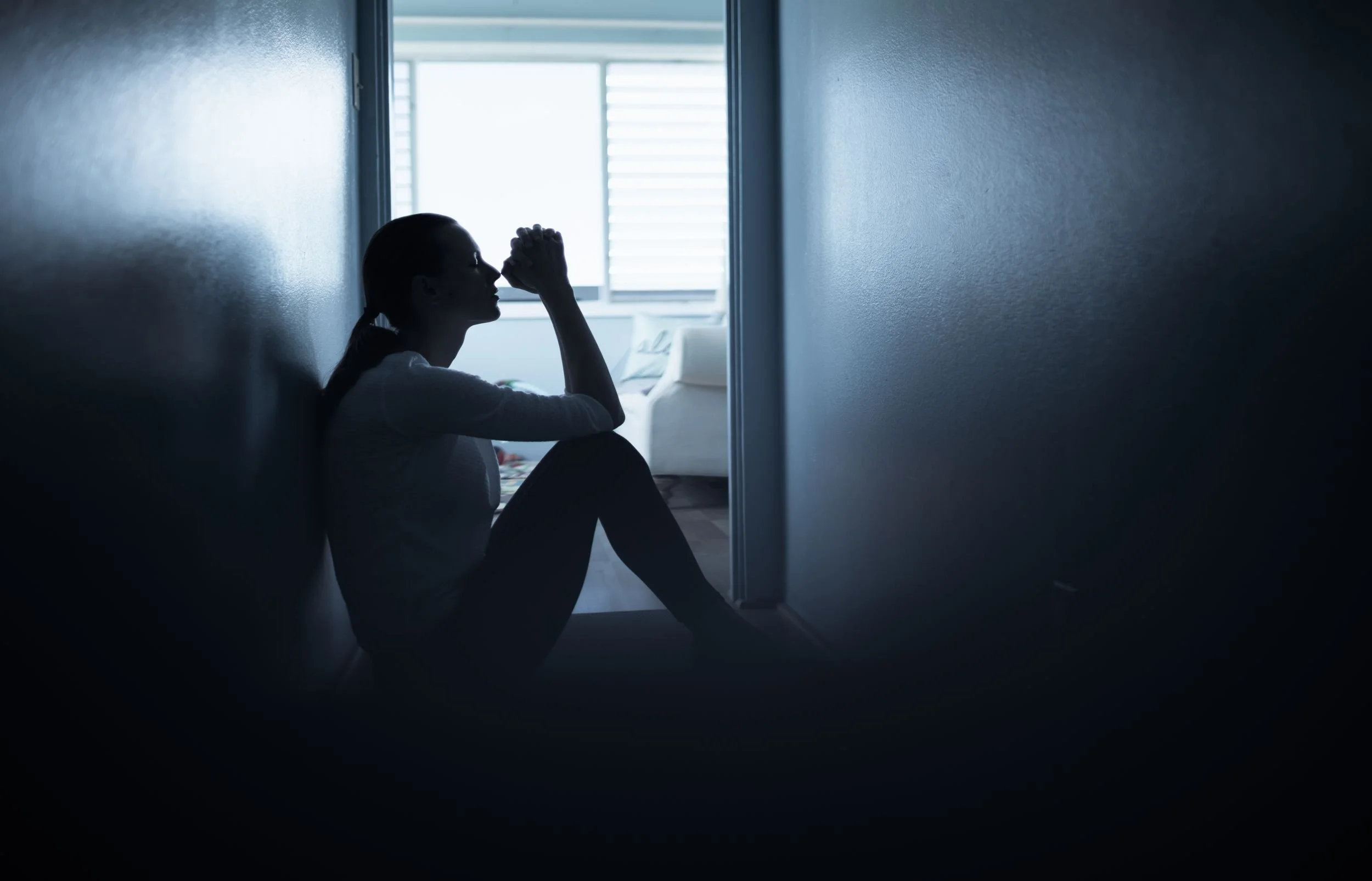 Eine Person sitzt in einem dunklen Raum, die Silhouette ist sichtbar, während sie mit geschlossenem Blick und einer Hand an der Stirn nachdenklich aussieht. Im Hintergrund ist ein hell erleuchtetes Fenster, das den Raum von außen beleuchtet.