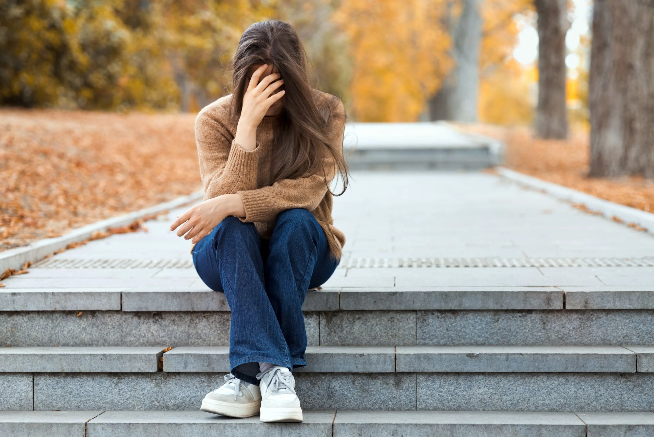 Junge Frau sitzt auf Treppenstufen in einem Park im Herbst, die Hand an den Kopf gelegt, mit bunten Blättern und Bäumen im Hintergrund.