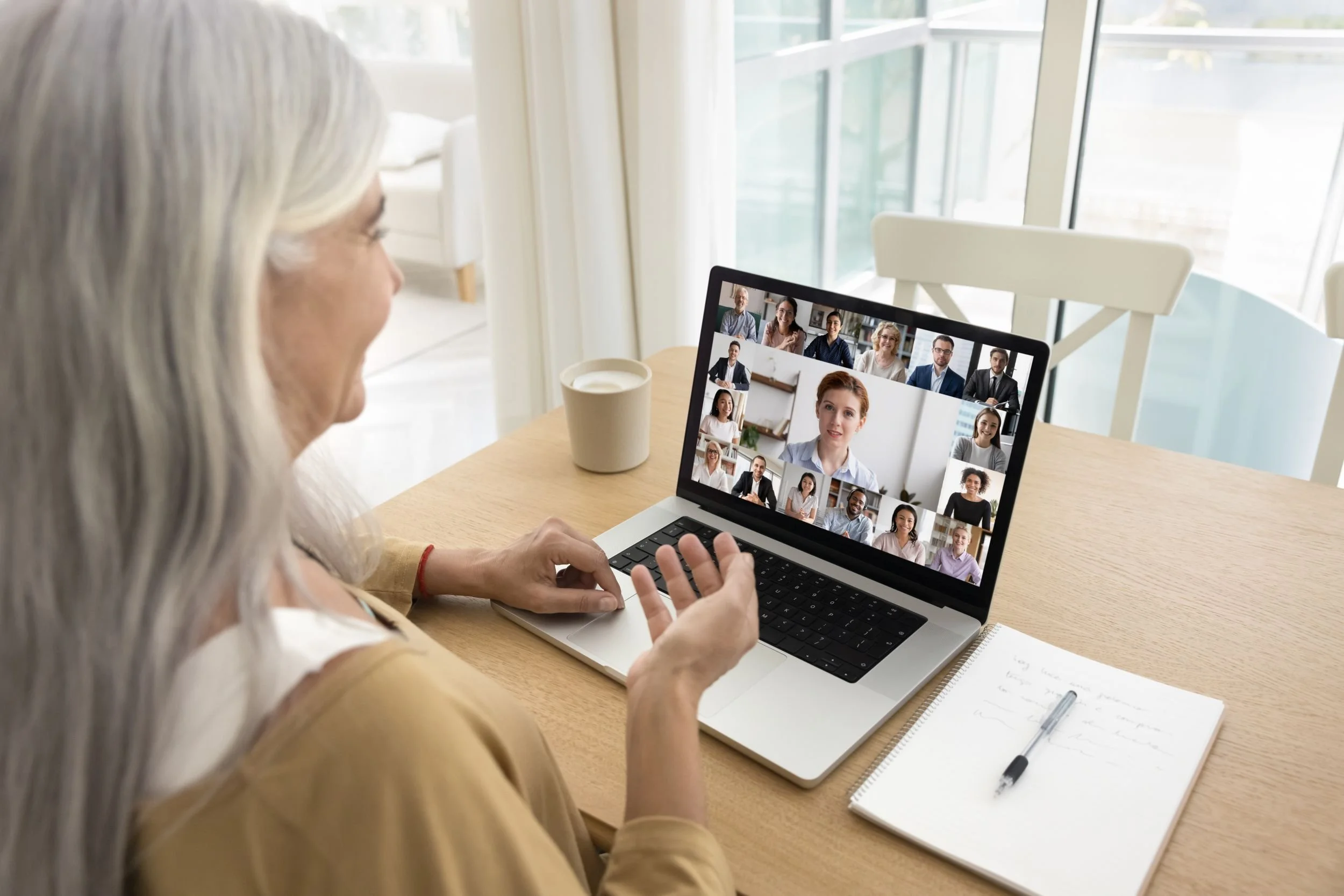 Ältere Frau nimmt an einem virtuellen Meeting auf einem Laptop in einem hellen Raum teil, mit mehreren Teilnehmern auf dem Bildschirm sichtbar.