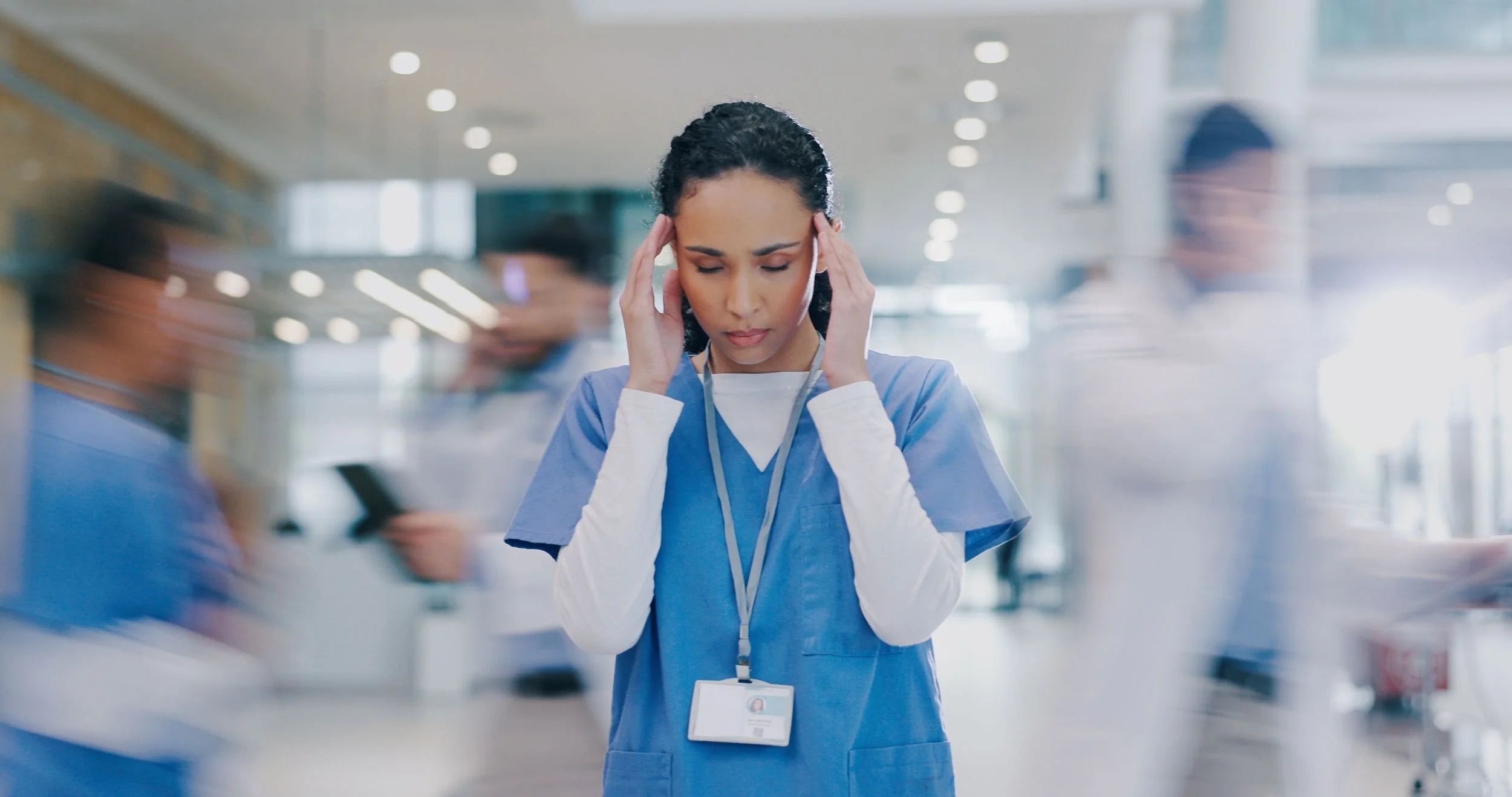 Eine Krankenschwester in blauer Arbeitskleidung hält sich die Schläfen und sieht besorgt aus, in einem Krankenhausflur mit anderen Personen im Hintergrund.