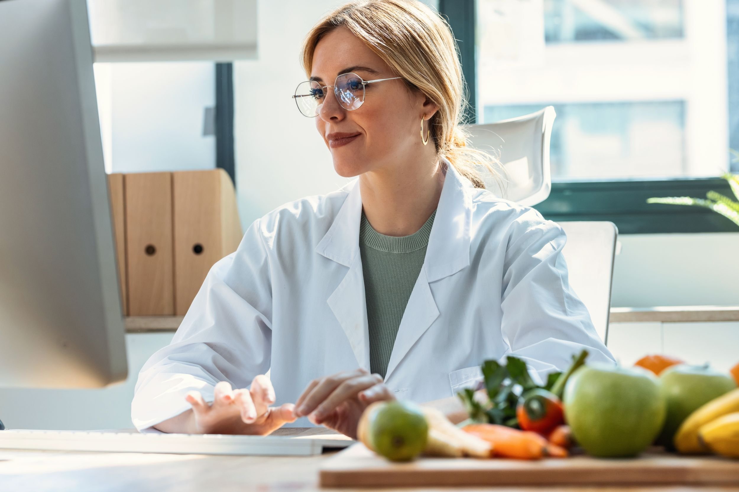 Eine Frau in einem weißen Laborkittel und Brille arbeitet in einer modernen Küche mit großen Fenstern und natürlichem Licht, umgeben von frischem Obst und Gemüse.