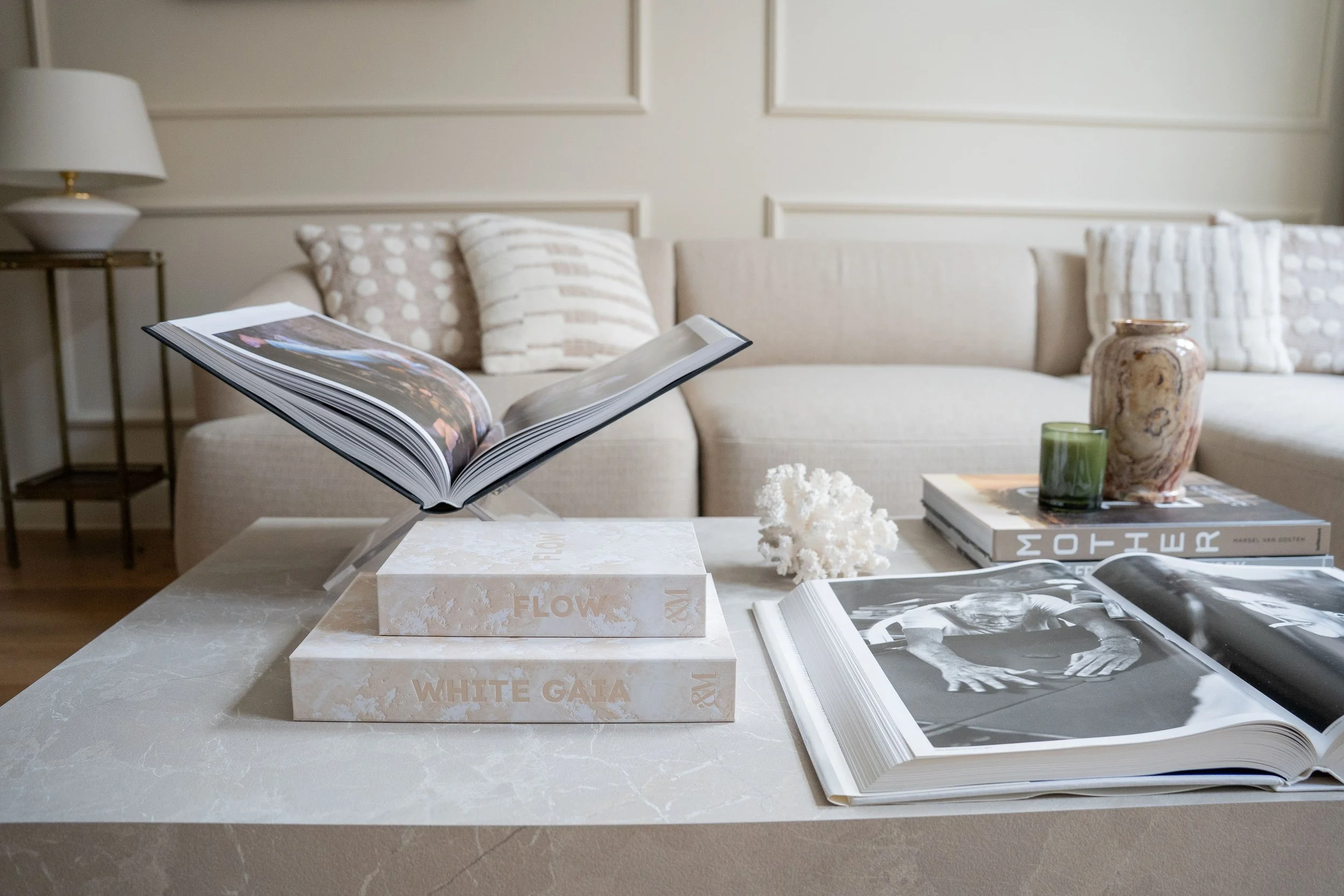 Hoge boeken met foto's en tekst liggen op een wit tafelblad in een woonkamer, met een bank en decoratieve items op de achtergrond.
