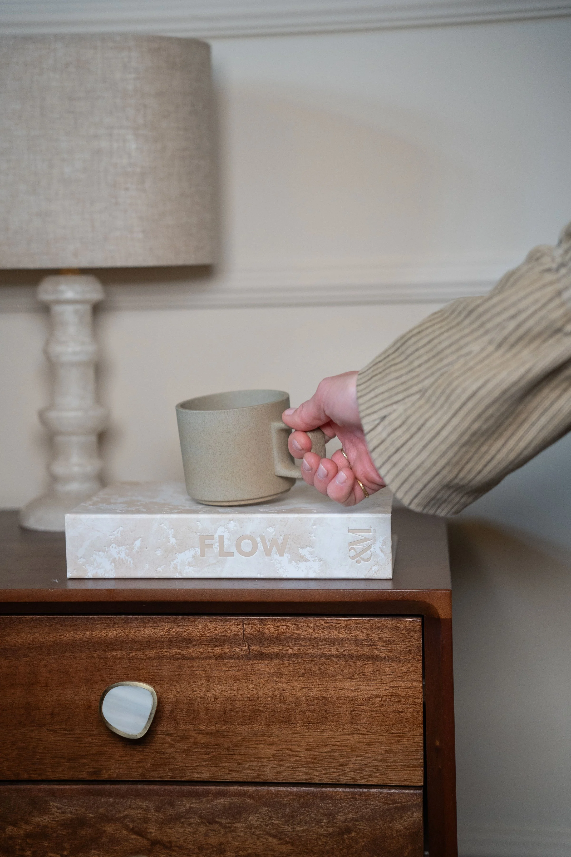 Een hand die een beige mok pakt van een opbergboek met de tekst 'FLOW' op een nachtkastje, naast een lamp en een spiegel.