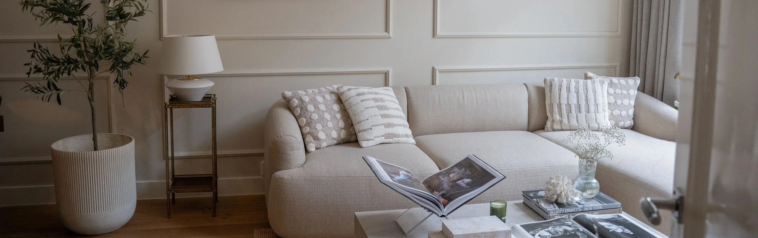 Een woonkamer met een beige L-vormige bank, versierd met vier beige en witte kussen, een salontafel met boeken, een plant in een grote witte pot en een Circulaire tafellamp van Indexandmore op een klein salontafeltje.