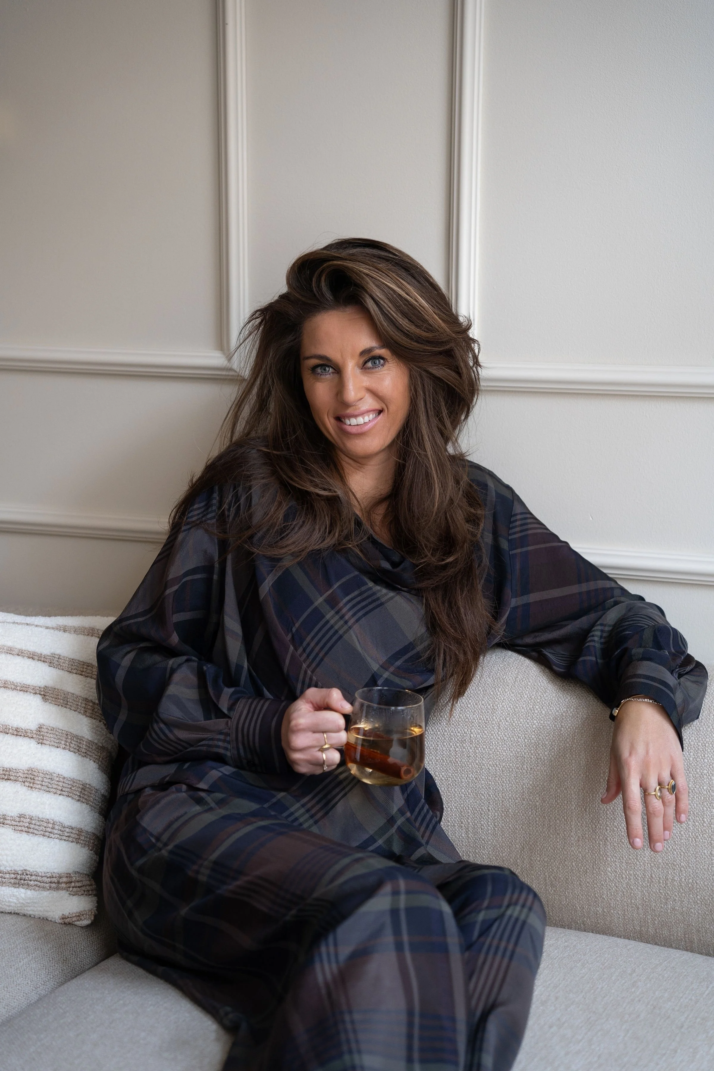 Vrouw met lang, bruin haar zit op een bank en houdt een glas thee vast.