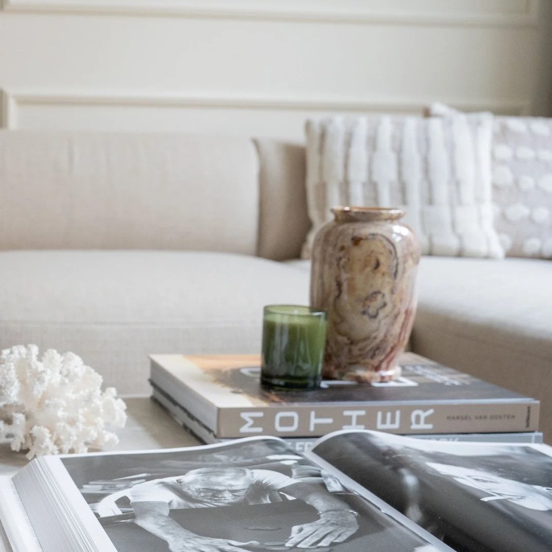 Een salontafel met een koffietafelboek, een  kaars, een vaas met een marmerpatroon uit "The Spotlight van Indexandmore"en decoratieve kussens op een bank in een woonkamer.