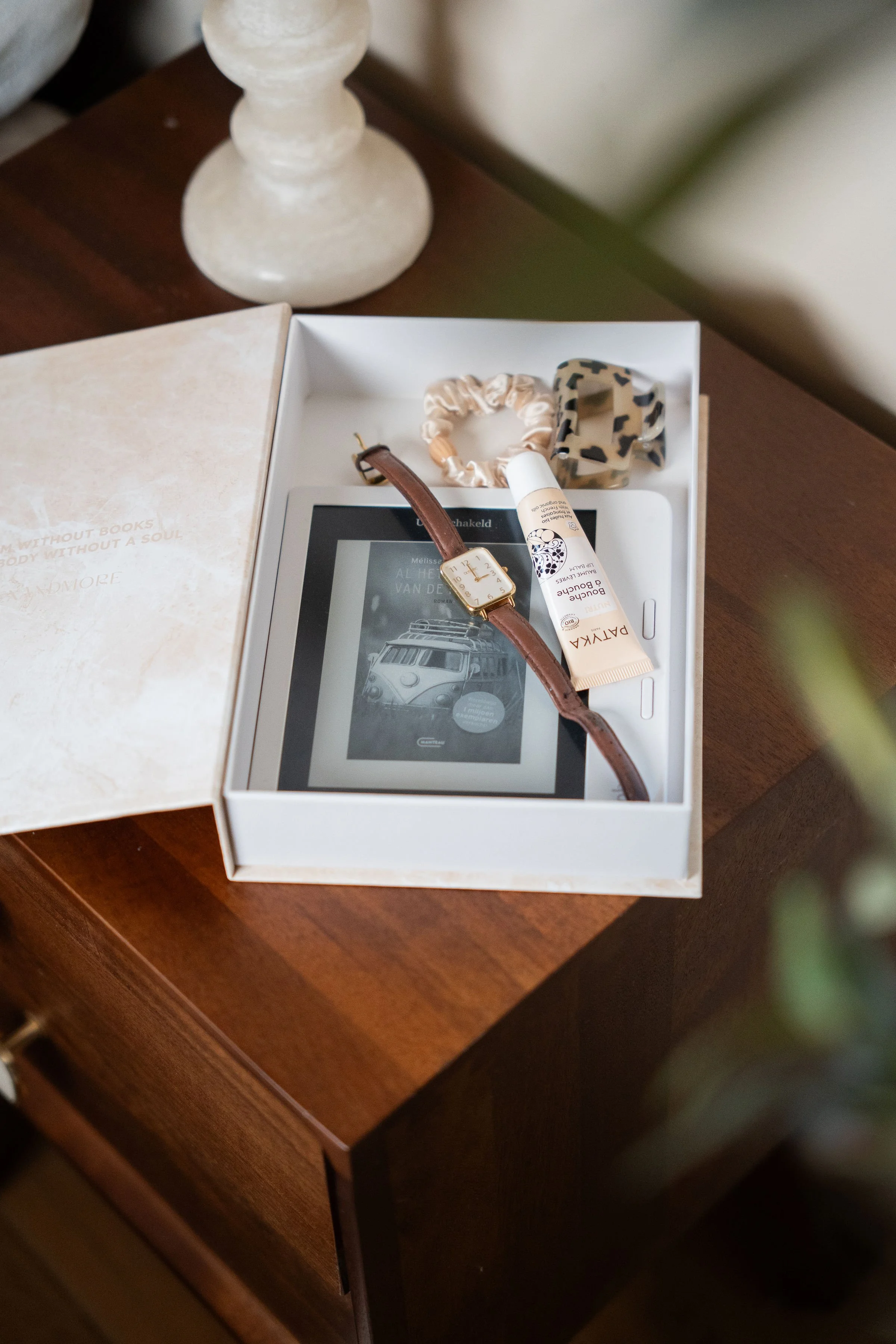 Indexandmore opbergboek ligt open met daarin: een e-reader, een horloge met een leren band, twee haarbanden en een tube huidverzorgingsproduct op een houten tafel.