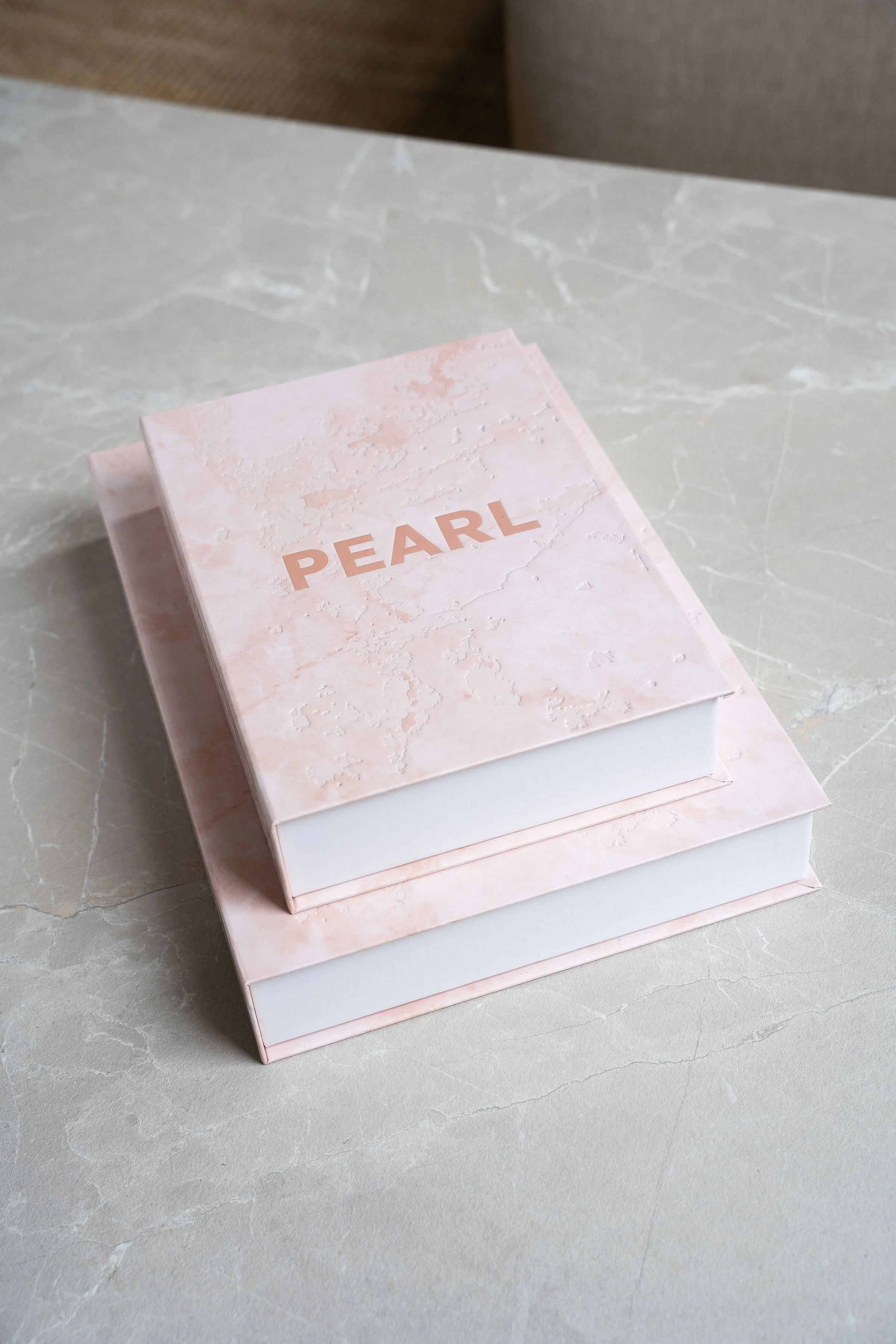 Een opbergboek genaamd 'PEARL' ligt op een marmeren tafel. Het boek heeft een roze, marmerachtig eiwitpatroon en ligt bovenop een ander opbergboek.