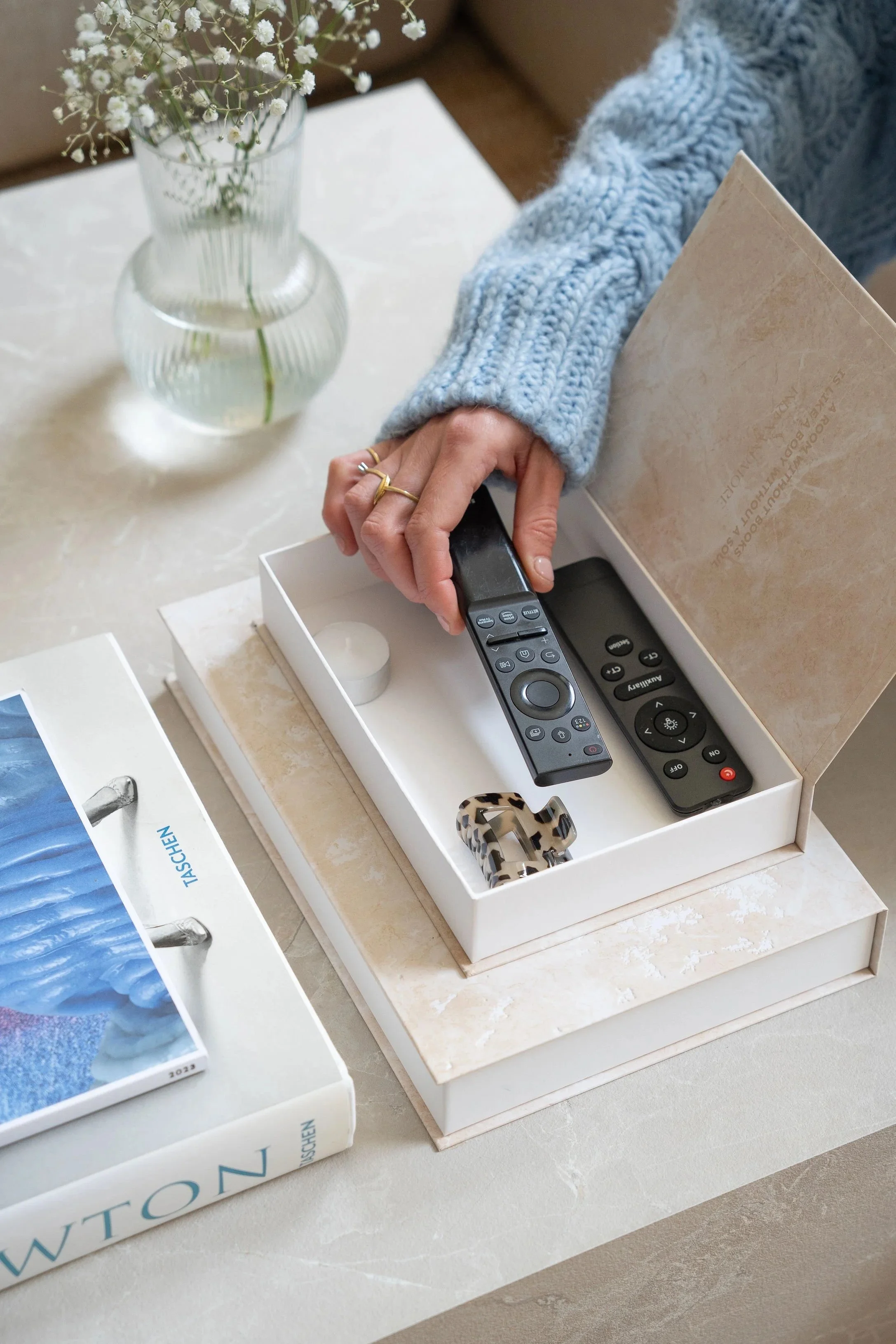 Een hand in een blauw gebreid wollen trui pakt een afstandsbediening uit een open opbergboek van Indexandmore. Op de tafel staan ook een boek en een vaas met kleine witte bloemen.