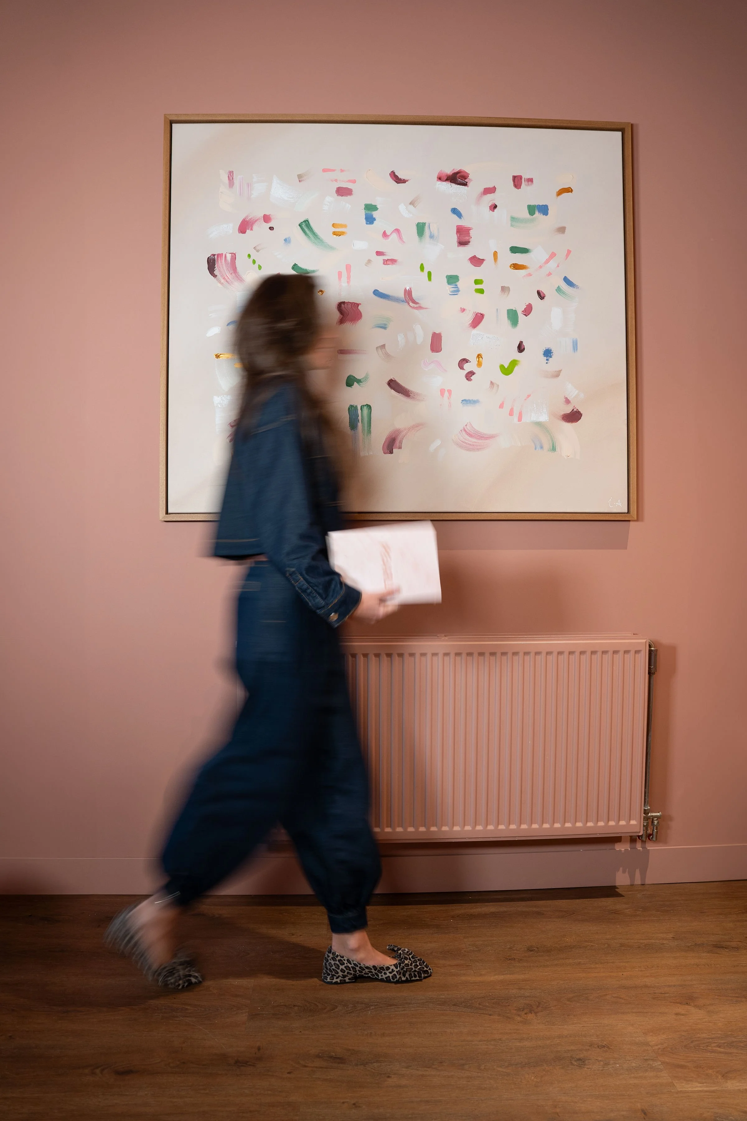 Een vrouw loopt langs een kunstwerk met een roze muur en houten vloer.