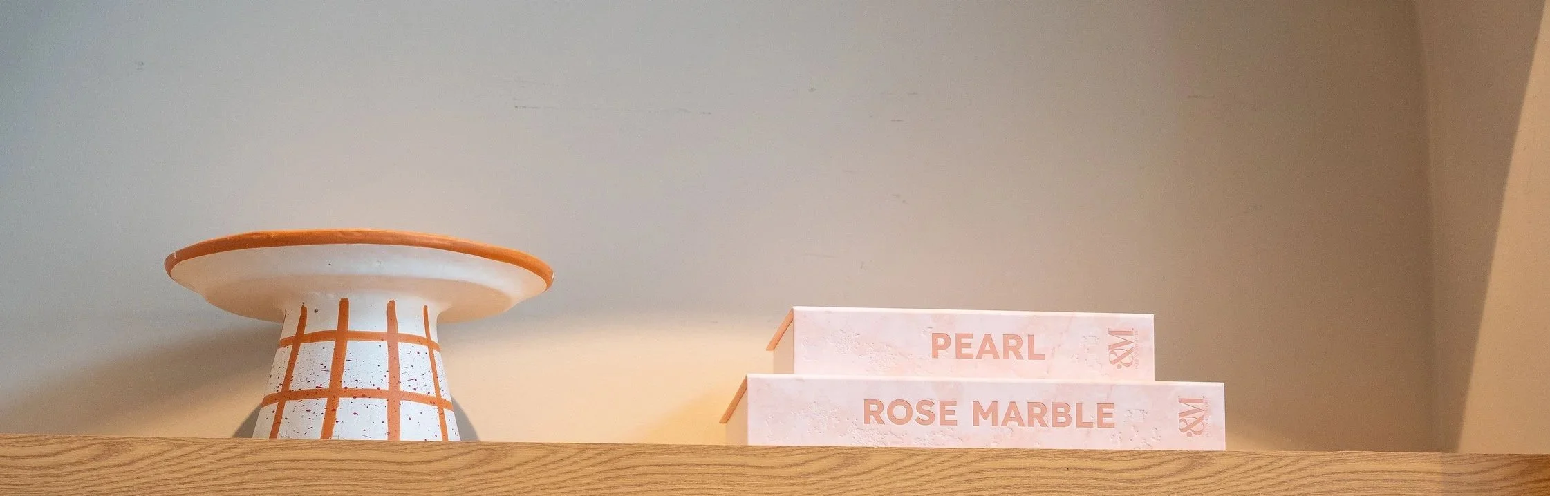 Decoratief wit en oranje dienblad op een houten plank, naast twee opbergboeken met roze marble covers getiteld 'Pearl' en 'Rose Marble'.
