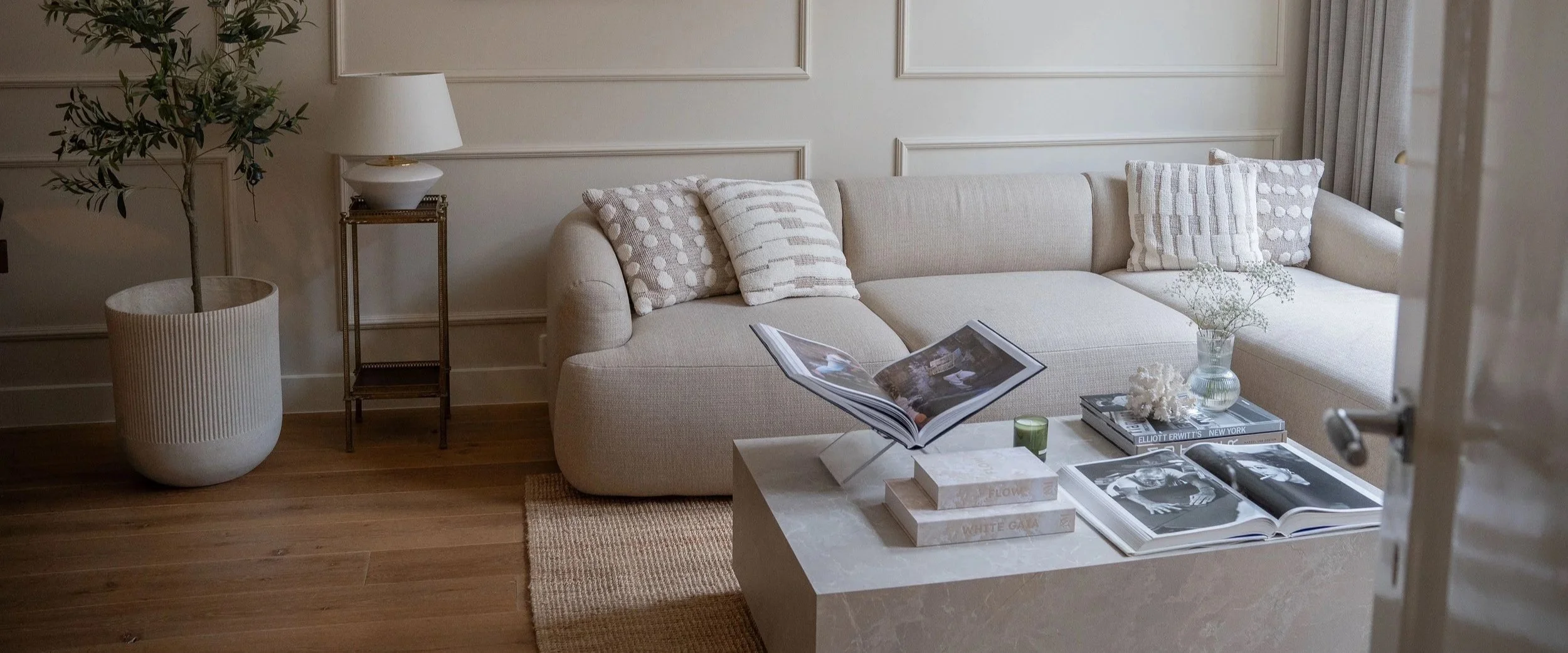 Gezellige woonkamer met witte bank, zachtgekleurde kussens, grote ramen met zandkleurige gordijnen, houten vloer, salontafel met decoratieve boeken, opbergboeken en accessoires.