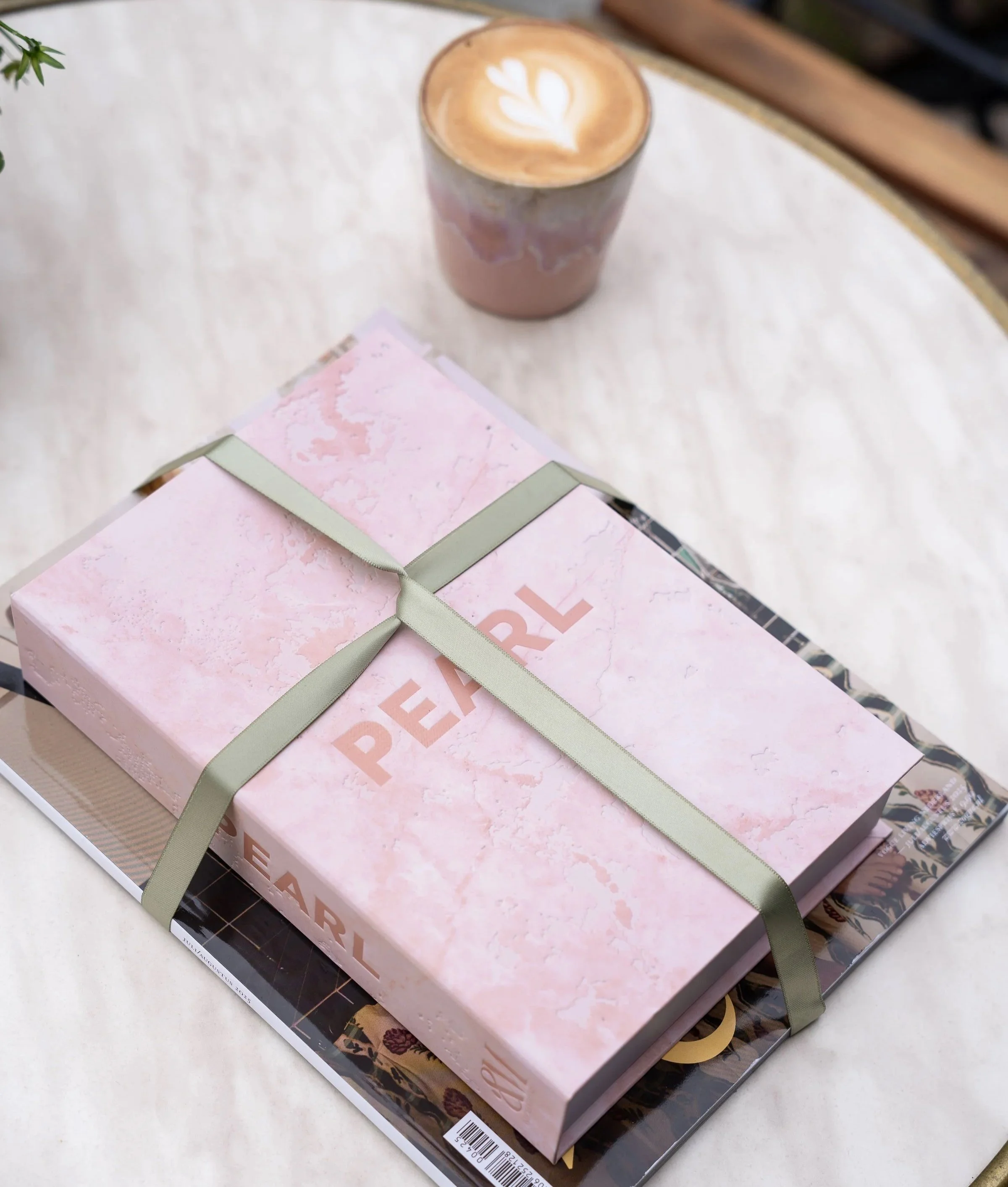 Een cadeau met een opbergboek met roze marmeren patroon en de tekst 'PEARL' erop, gebonden met een lichtgroene lint, ligt op een magazine op een tafel. Naast het cadeau staat een kop koffie met latte art.